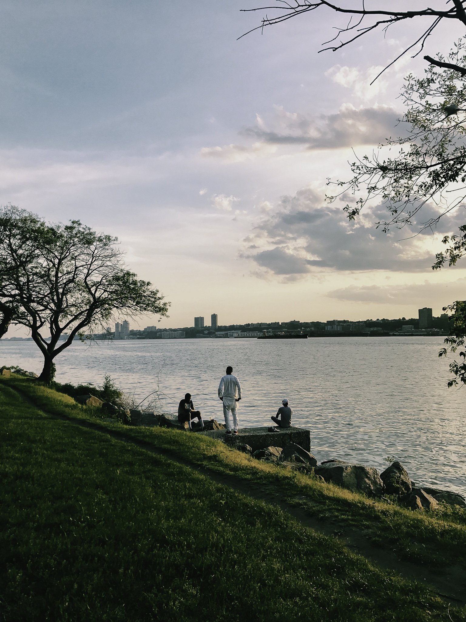 Three people sitting and standing on rocks by a river at sunset, with city buildings in the distance and a grassy area with trees in the foreground.