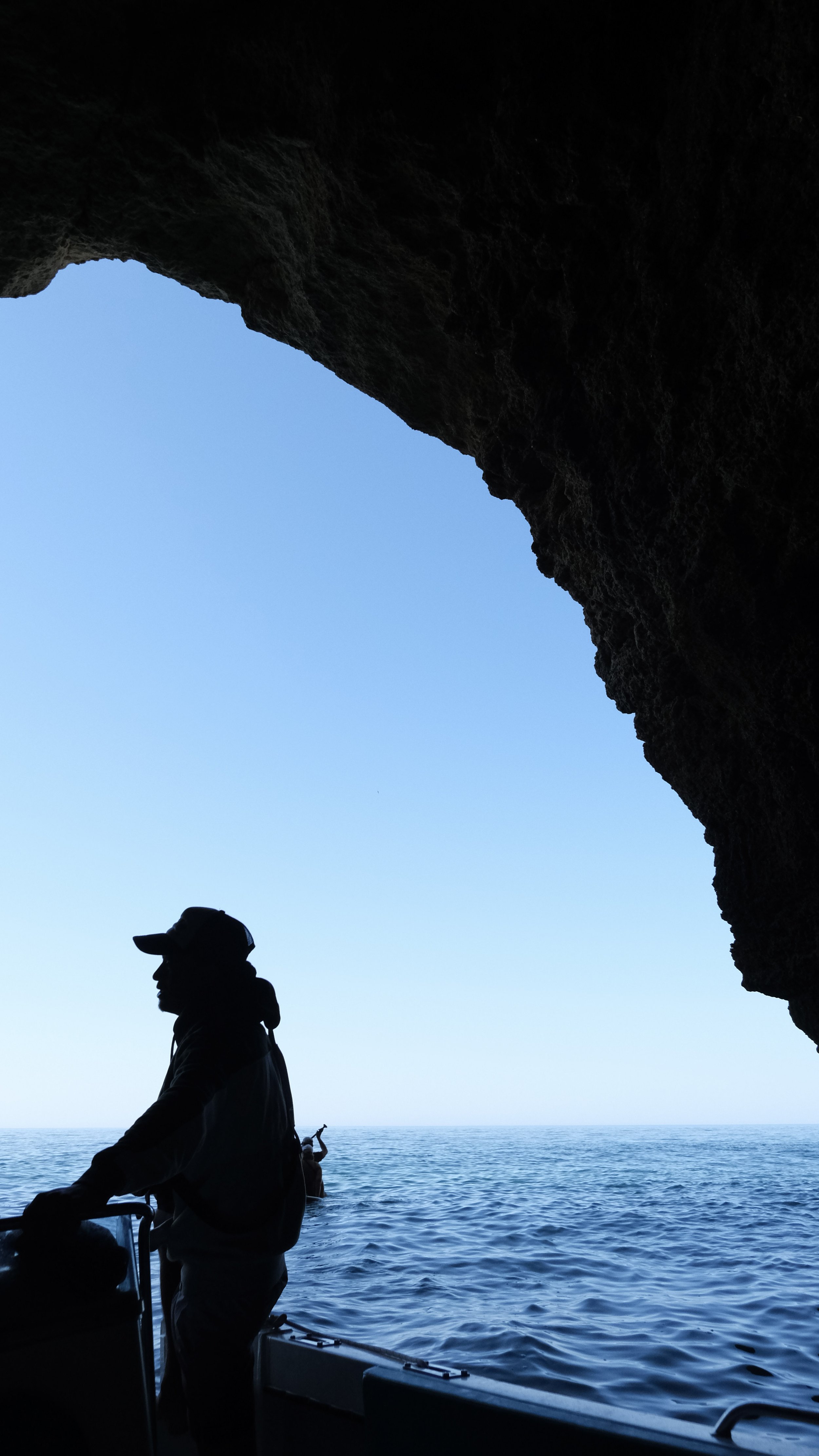 Silhouette of a person on a boat at sea, viewed from inside a cave or rock formation, with a clear blue sky and calm ocean in the background.