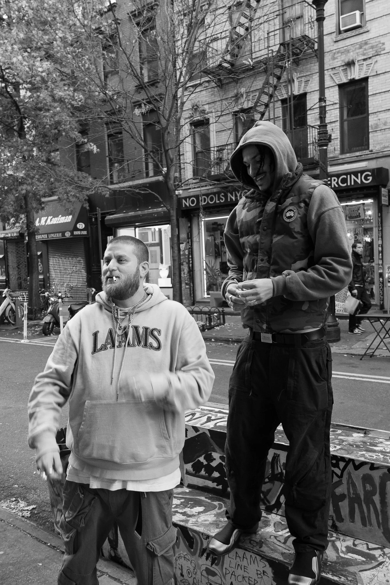 Two men standing on a city street sidewalk, one with a beard and hoodie, the other with a hooded jacket and ankle monitor, in an urban neighborhood with storefronts and trees in the background.