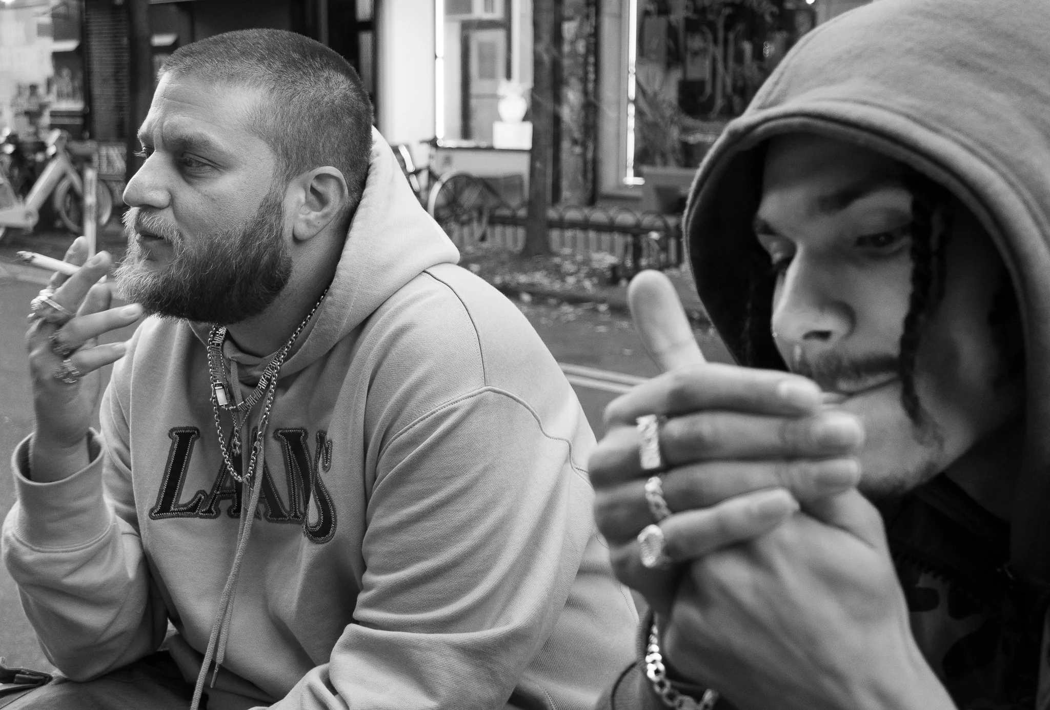 Two men sitting at a table outside, one with a beard and tattoos smoking a cigarette, the other with dreadlocks and wearing a hoodie, both looking down.