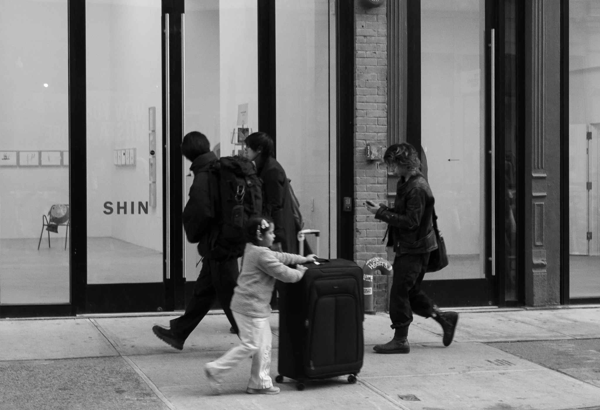 People walking past a glass storefront, including a woman and a young girl pushing a suitcase, and another woman looking at her phone.