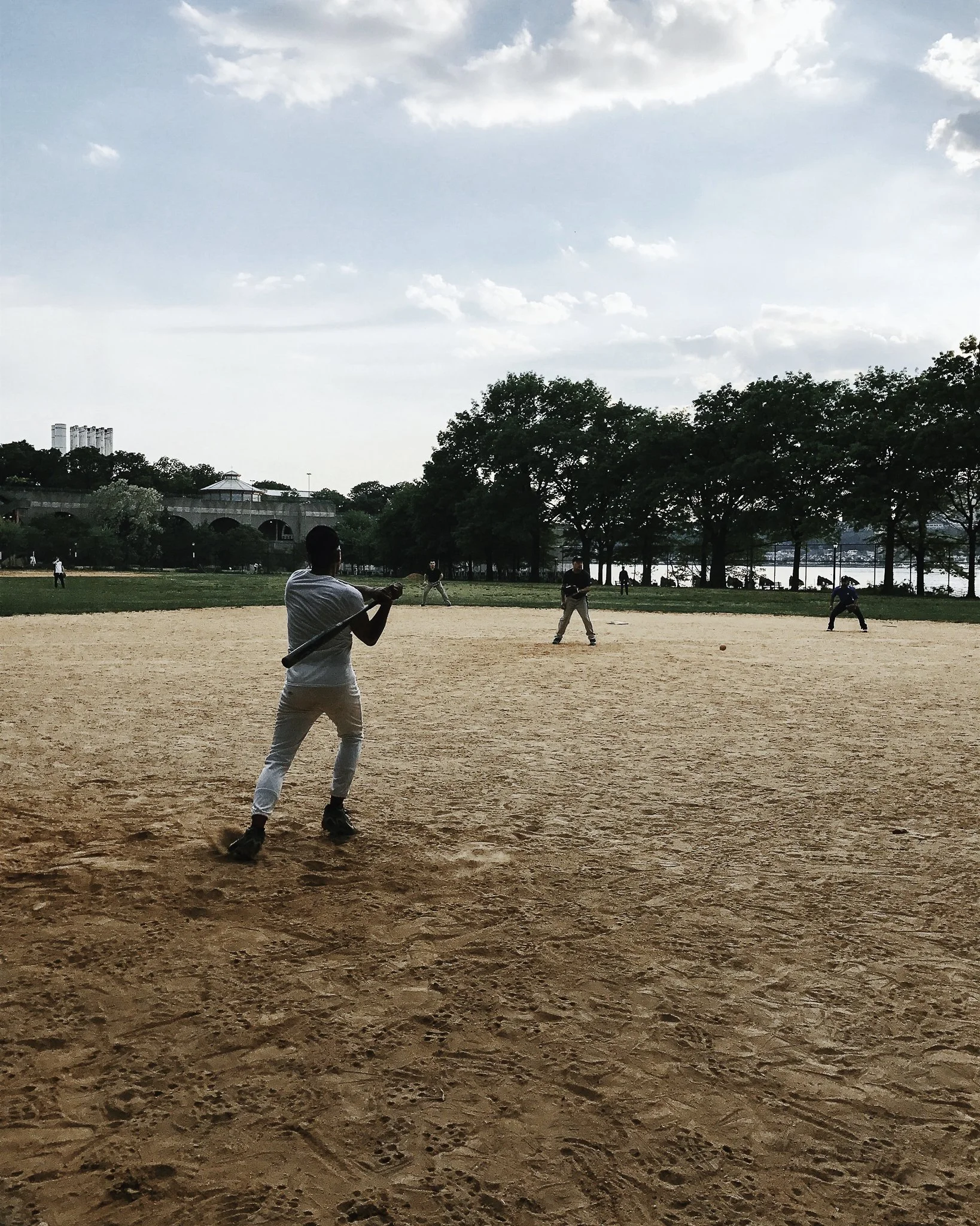 People playing baseball on a dirt field in a park with trees and a body of water in the background.