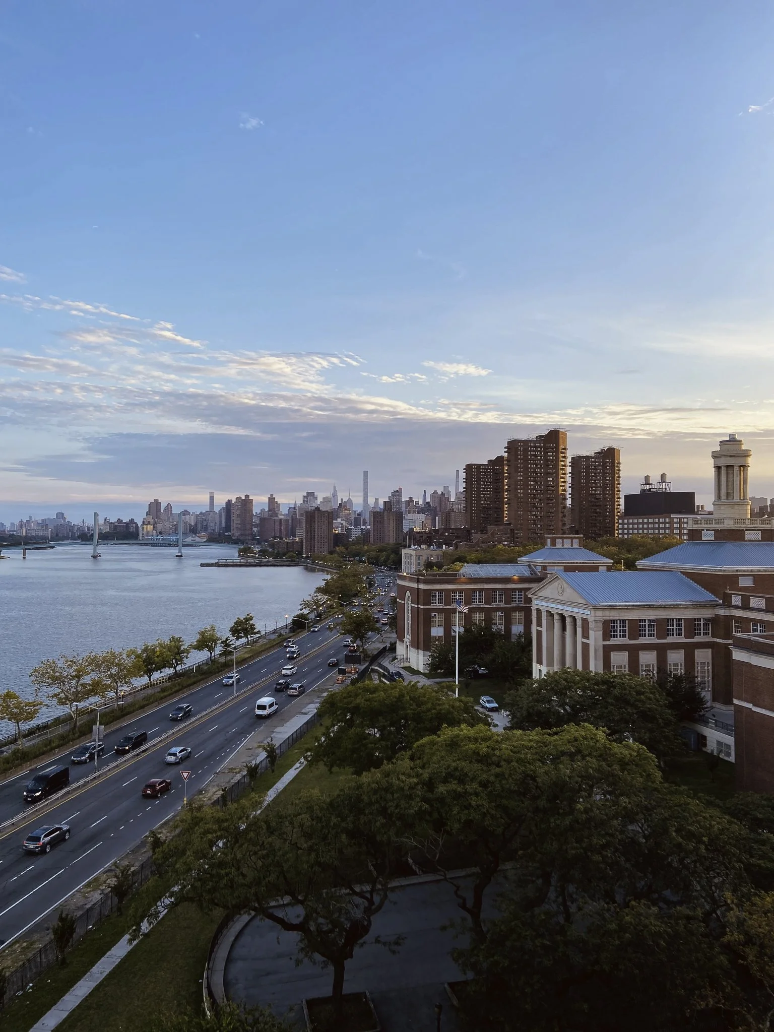 A city skyline near a body of water, with high-rise buildings, a bridge, a road with cars, and a park with trees, under a blue sky with some clouds.