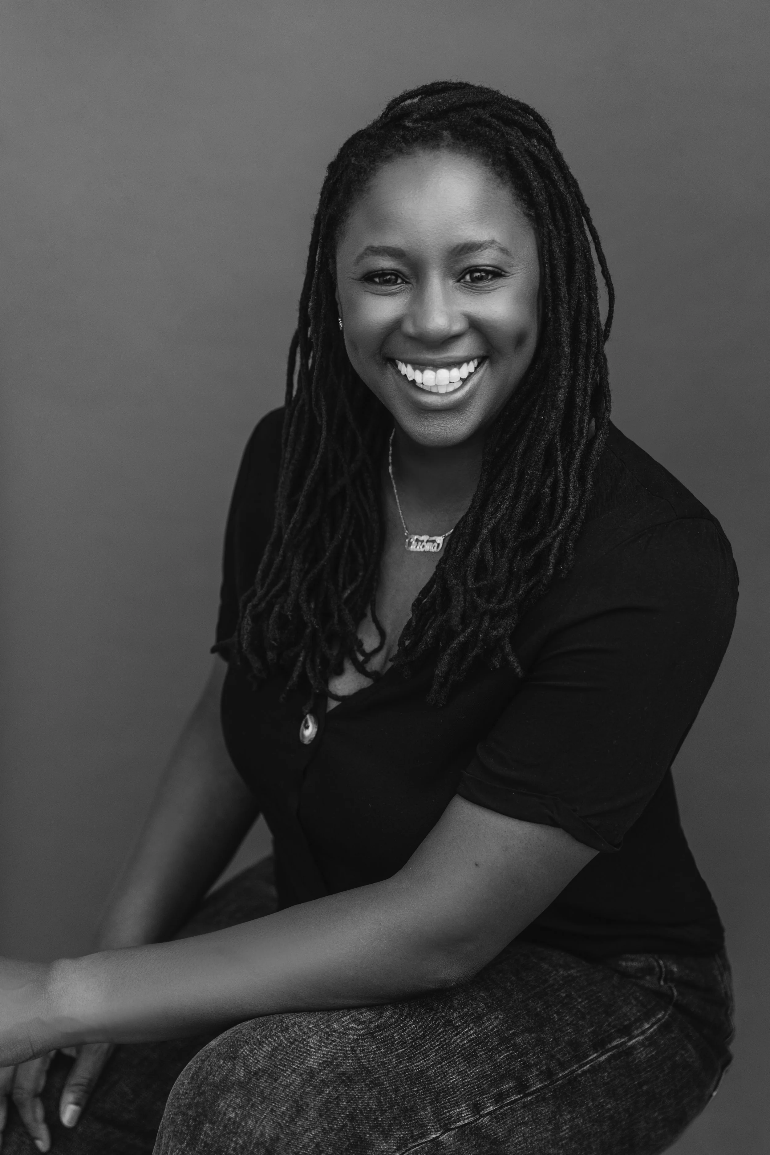 A black and white portrait of a woman smiling, with long dreadlocks, wearing a black top and jeans, sitting against a plain background.
