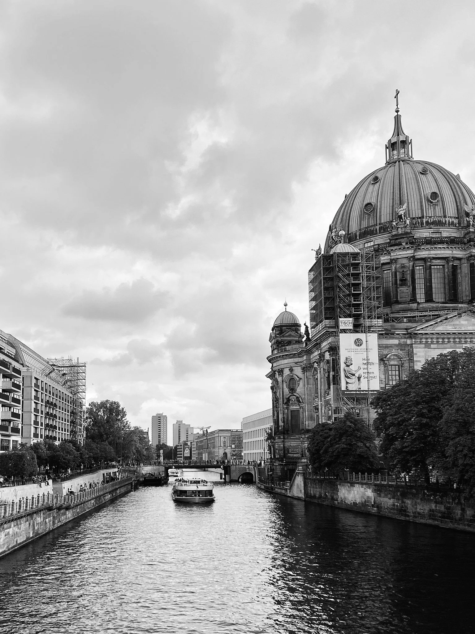 Black and white photo of the Berlin Cathedral with a canal and waterway in the foreground, boats on the water, trees lining the canal, and modern buildings in the background.