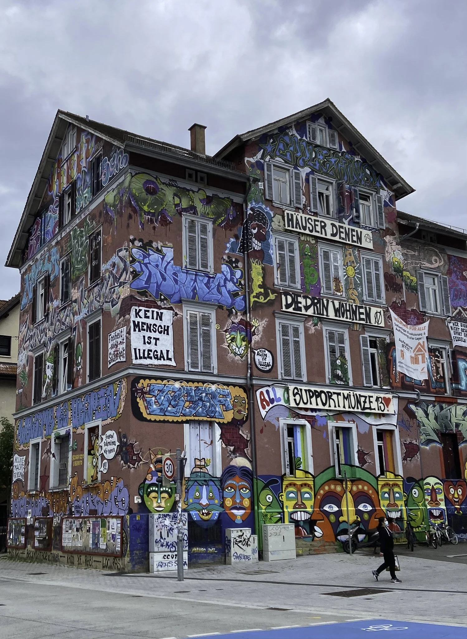 Colorful graffiti artwork covers a large multi-story building with various faces, symbols, and slogans in German.