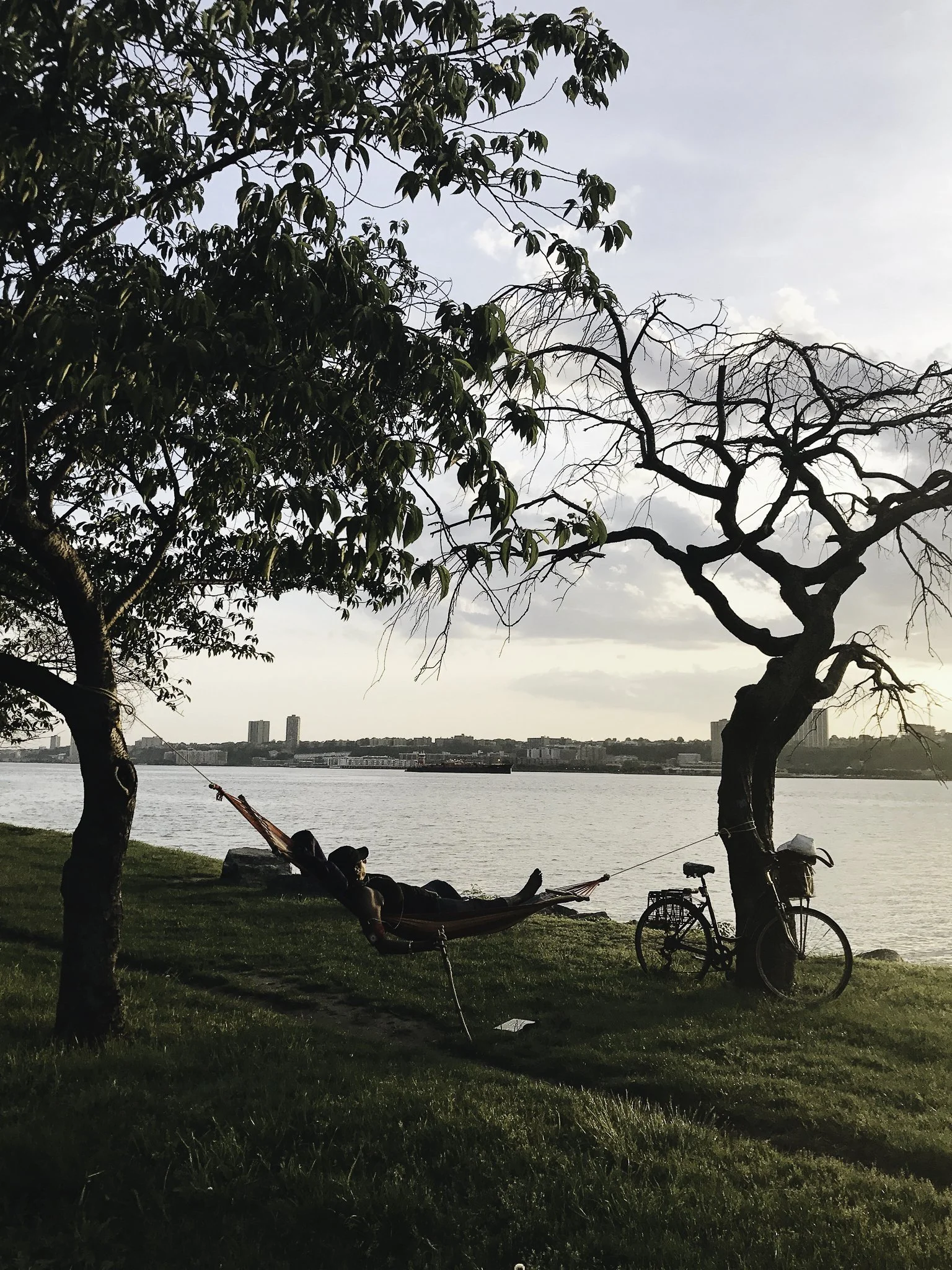 Person relaxing on a hammock tied between two trees by a river, with a bicycle leaning against one tree, in a park at sunset.