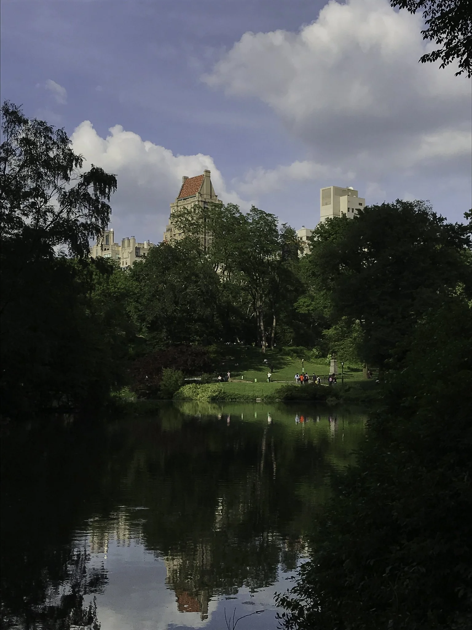 A park with a pond in the foreground, people walking and relaxing on the grassy hill, surrounded by trees, with buildings visible in the background under a partly cloudy sky.