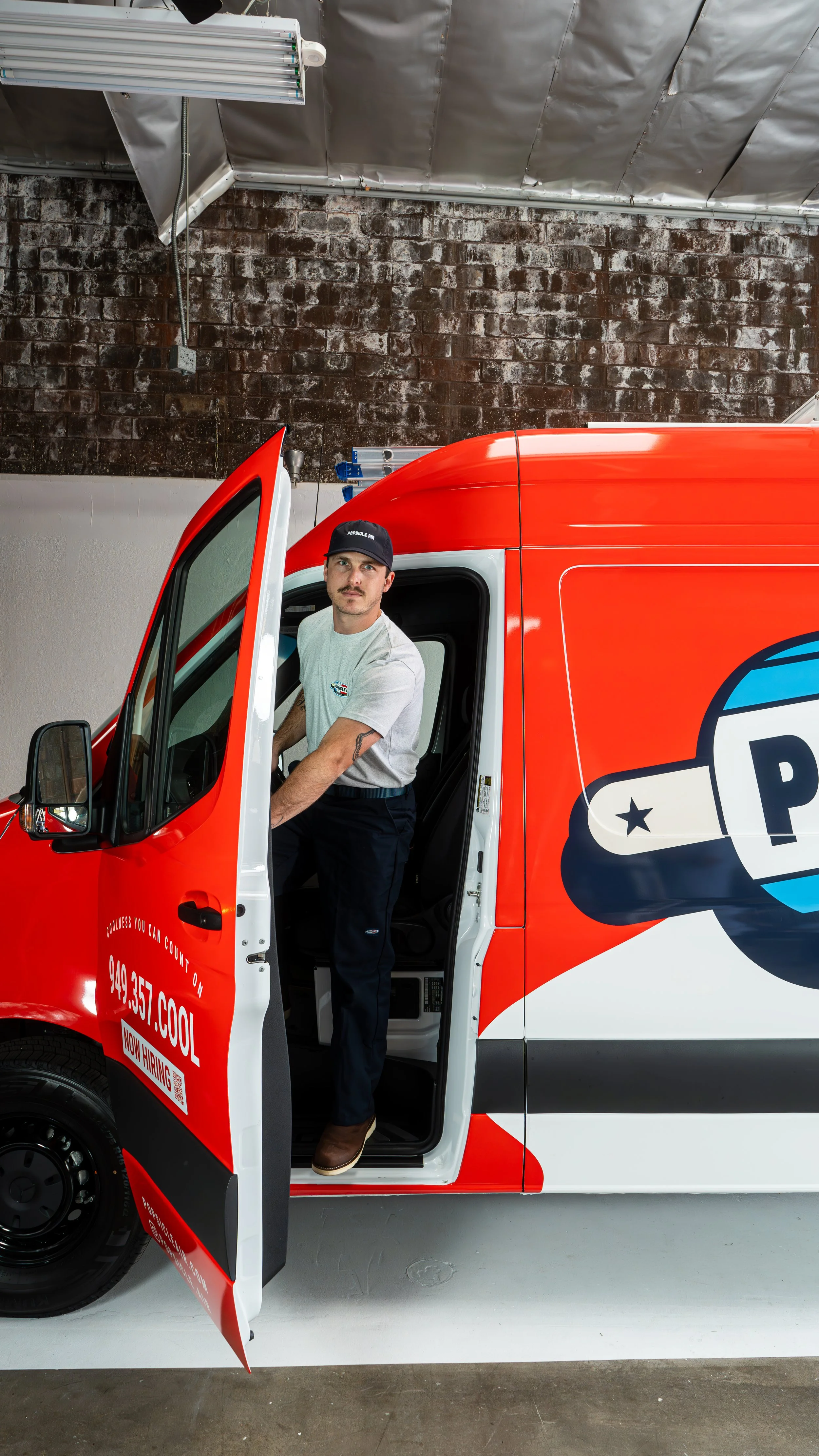 Person standing in the doorway of a red and white service van, wearing a uniform with a cap, in a garage or warehouse setting.
