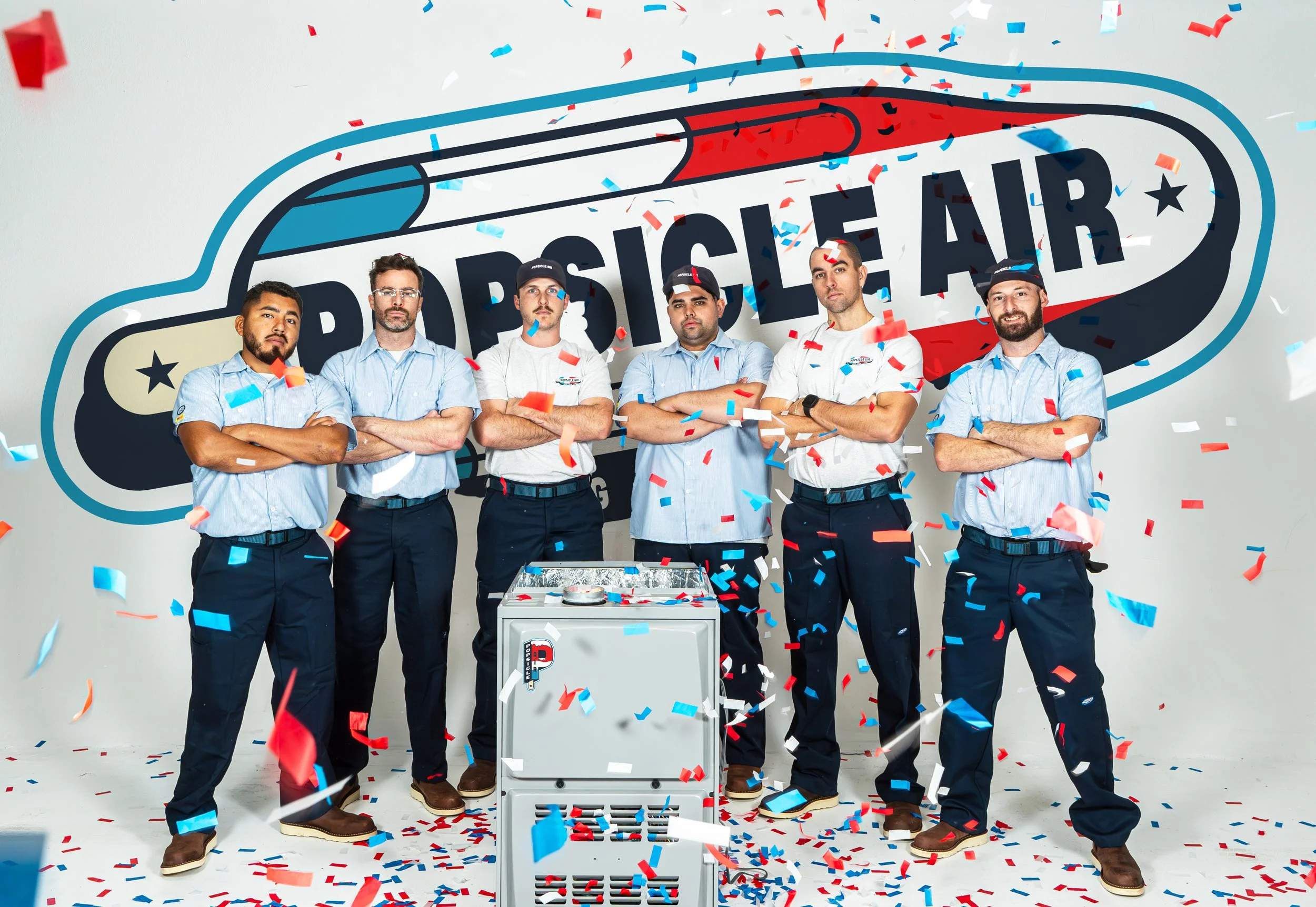 Group of six men in uniform posing with a Popsicle Air branding backdrop, surrounded by red and blue confetti.