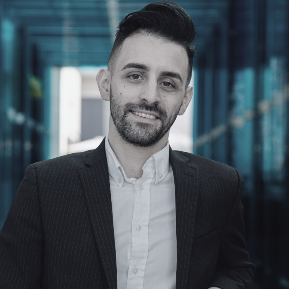 Michael Brito; A young man with dark hair, beard, and a friendly smile, wearing a white shirt and black blazer, standing outdoors in front of a modern glass building.