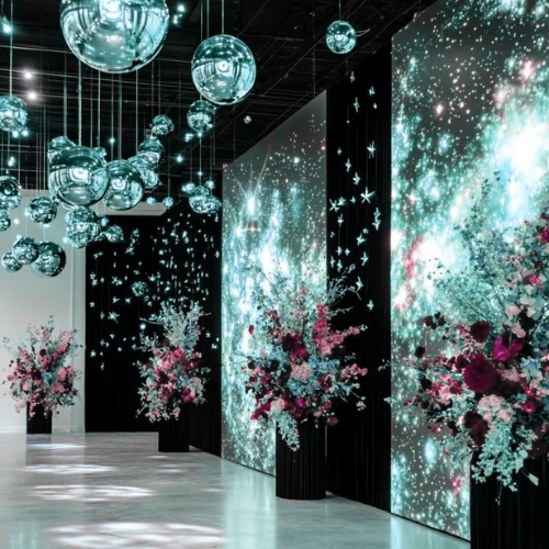 An art installation with hanging reflective spheres, large digital screens displaying starry night skies and cosmic patterns, and floral arrangements with pink and white flowers in black vases.