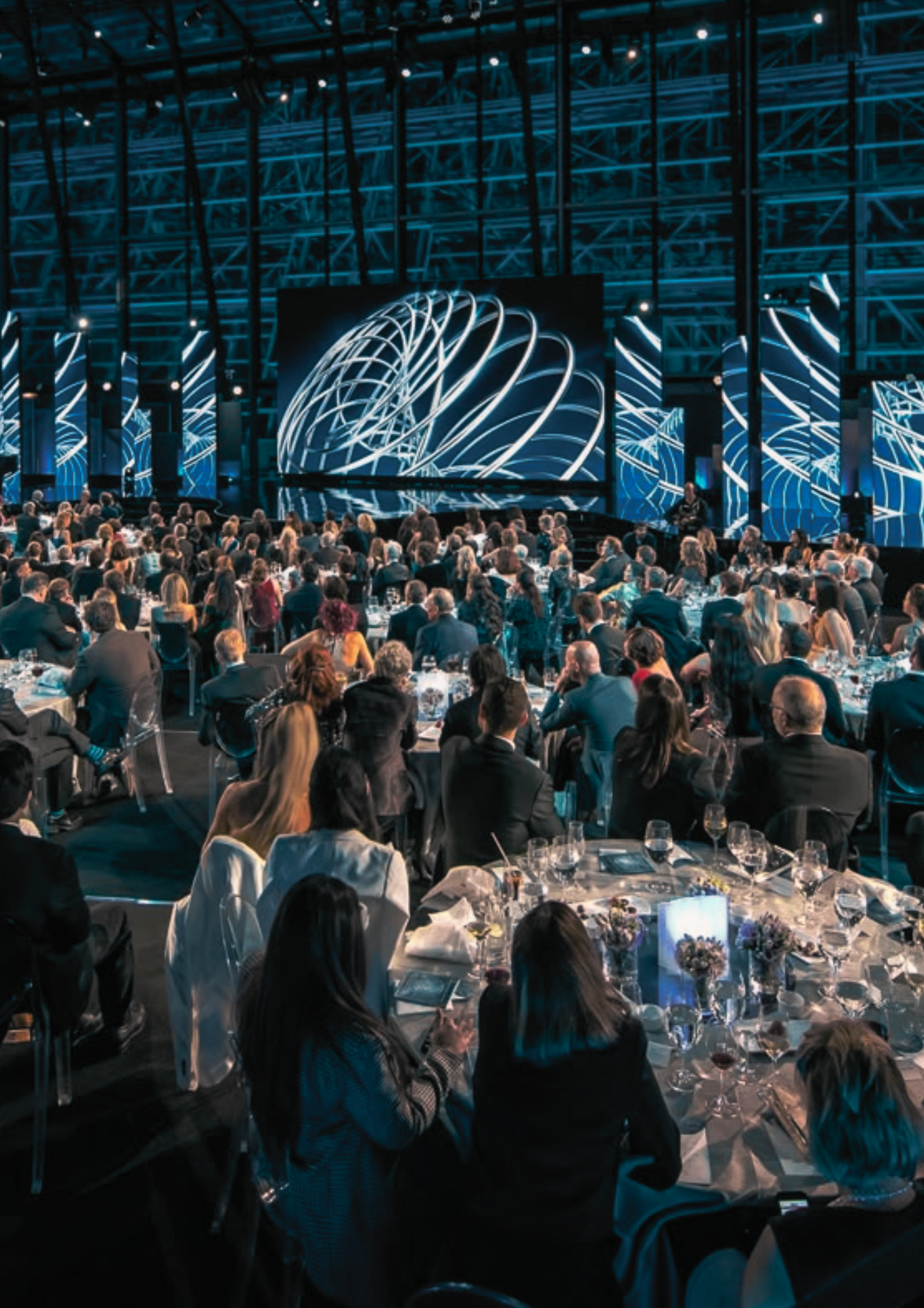Large conference or awards ceremony with many attendees seated at round tables, stage with large screen displaying abstract graphic, dark industrial-style venue with blue lighting.