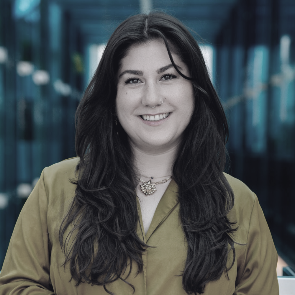 Danay Korsheh; A young woman with long, dark wavy hair smiling at camera, wearing a tan jacket and a floral necklace, standing outdoors in front of a modern glass building.