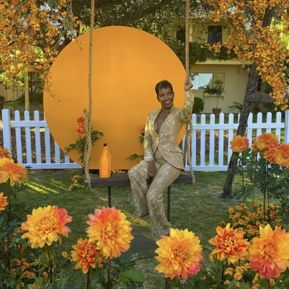 A woman in a gold-patterned suit sitting on a swing surrounded by orange and yellow flowers with a yellow backdrop and white picket fence in a garden.