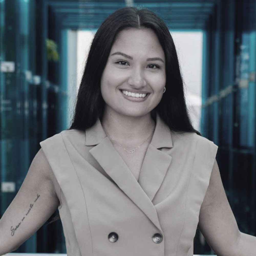 Stephanie Zuluaga; A woman with long dark hair smiling in a beige sleeveless blazer, standing outdoors in front of a modern glass building.