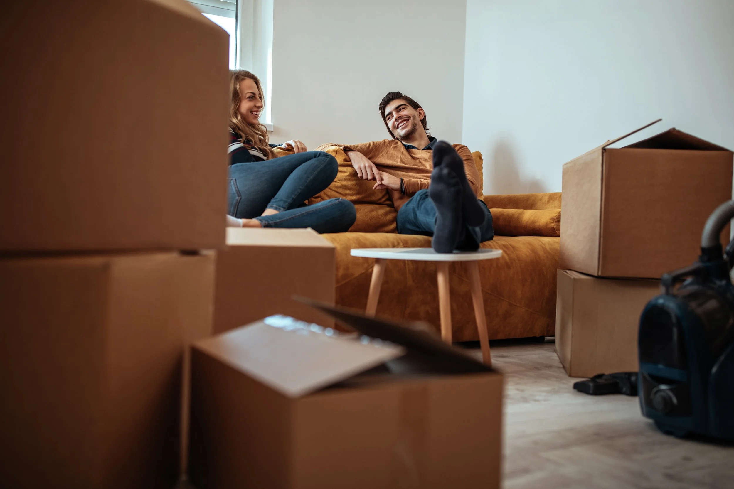 Couple sitting on their lounge smiling surrounded by moving boxes.