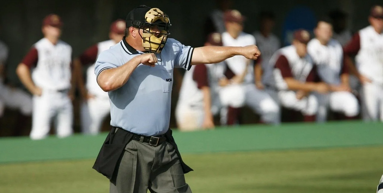 Umpire throwing a baseball in a game with players on the bench in the background.