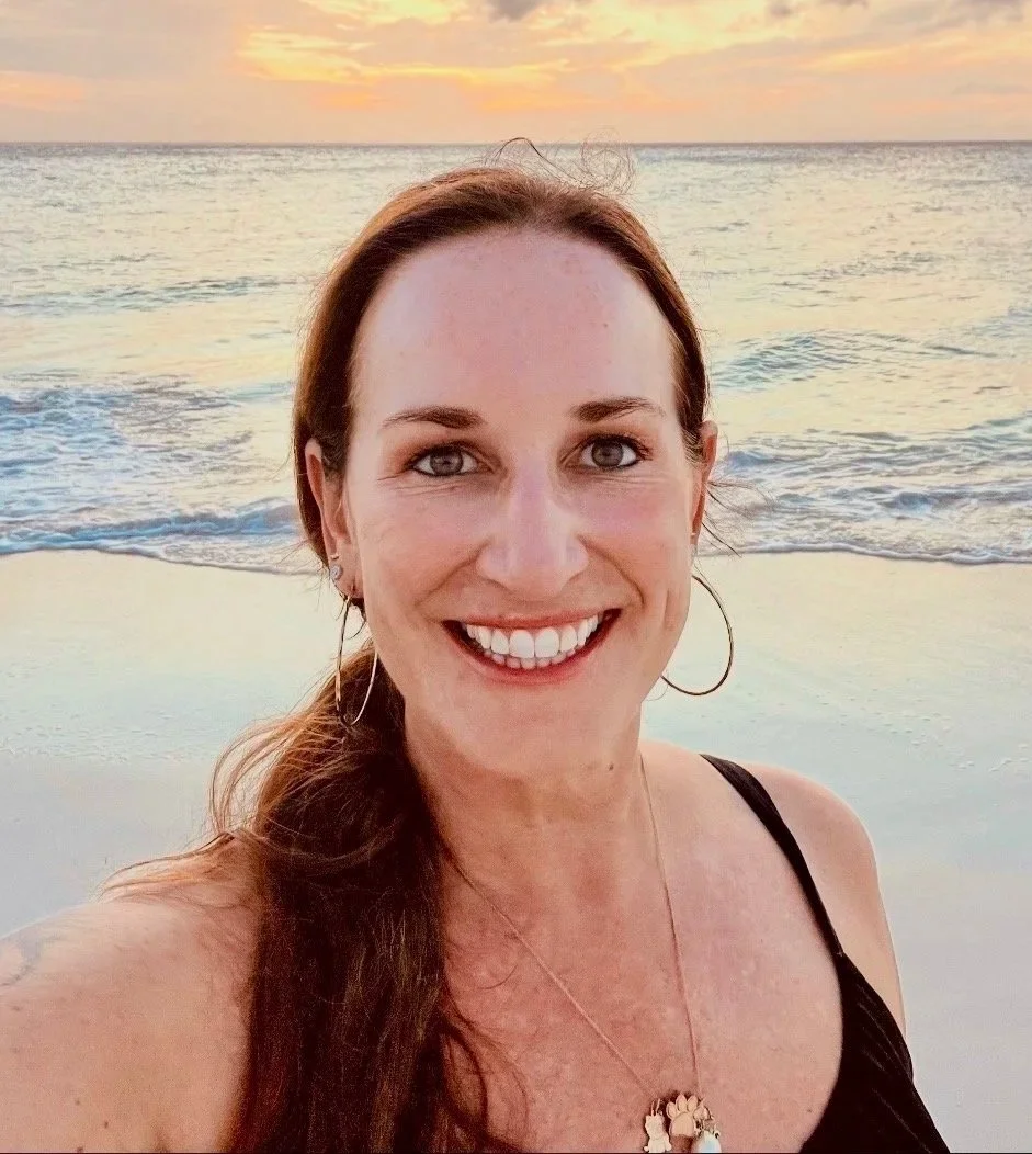 A woman smiling at the beach during sunset, with the ocean and sky in the background.