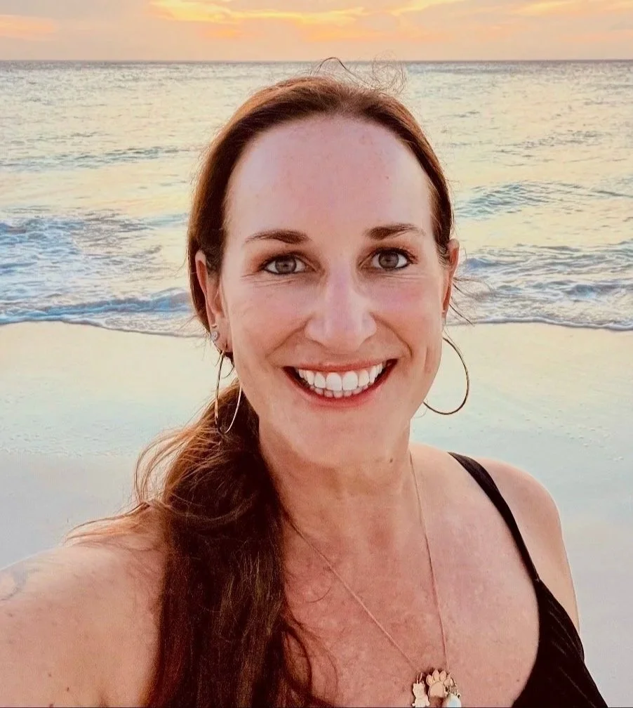 A woman taking a selfie on the beach during sunset, with the ocean and sky in the background.