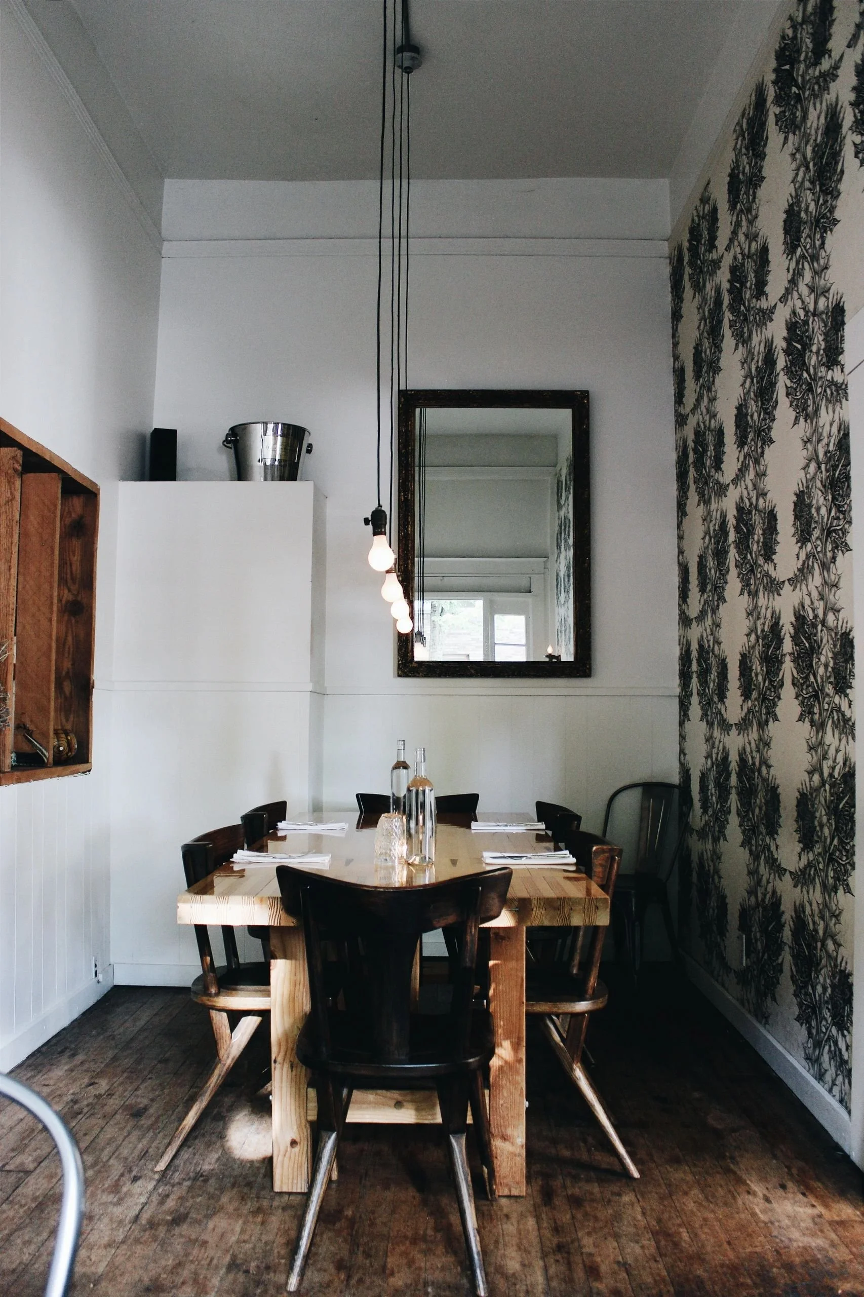 A dining room with a wooden table set for four, with water bottles and glasses. There are black chairs, one gray chair, a large mirror on the wall, hanging light bulbs, and wallpaper with a black and white botanical pattern on one wall.
