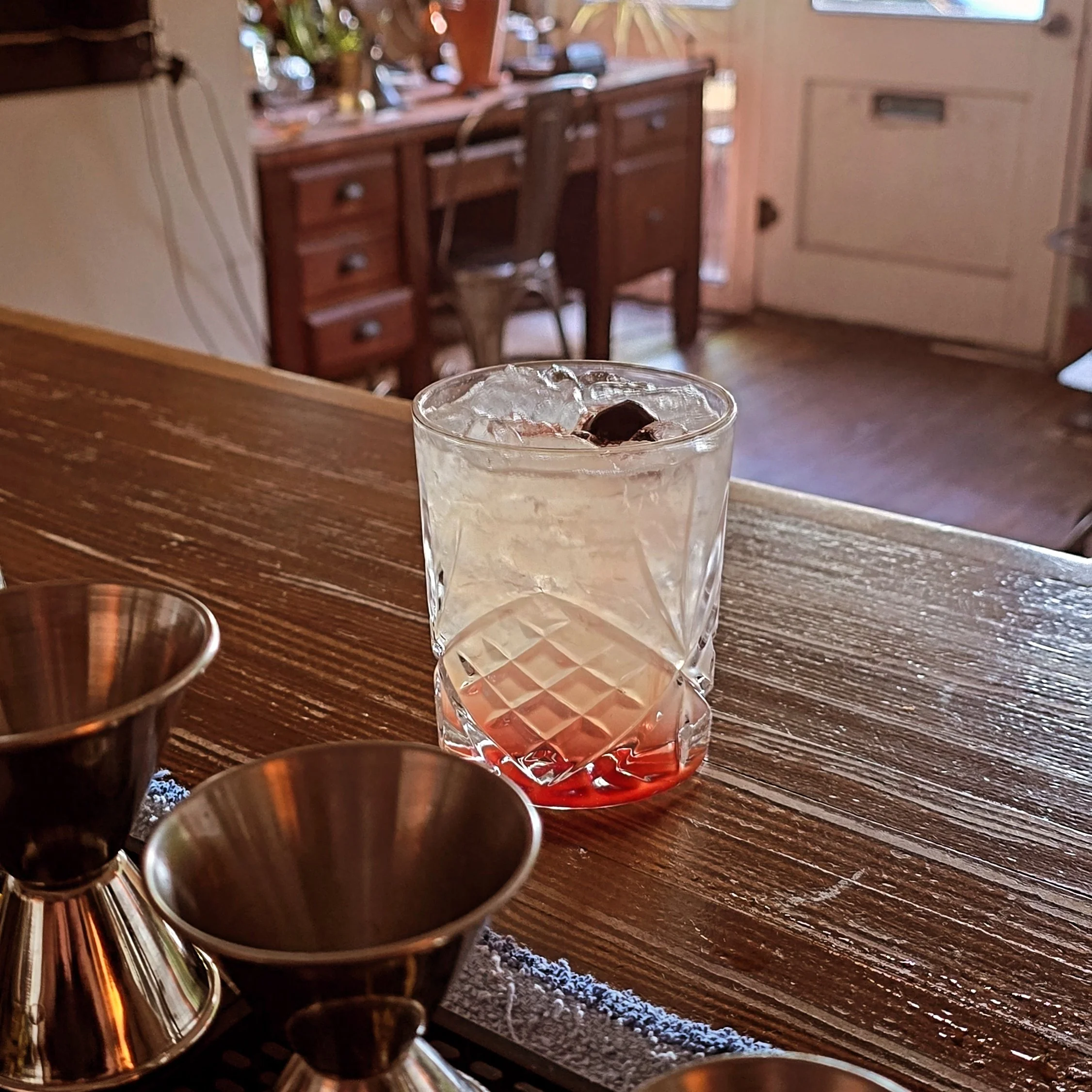 A glass of a cocktail with ice and a cherry on top on a wooden bar counter, with two metallic jiggers in front and a blurred background of a cozy kitchen or bar area.