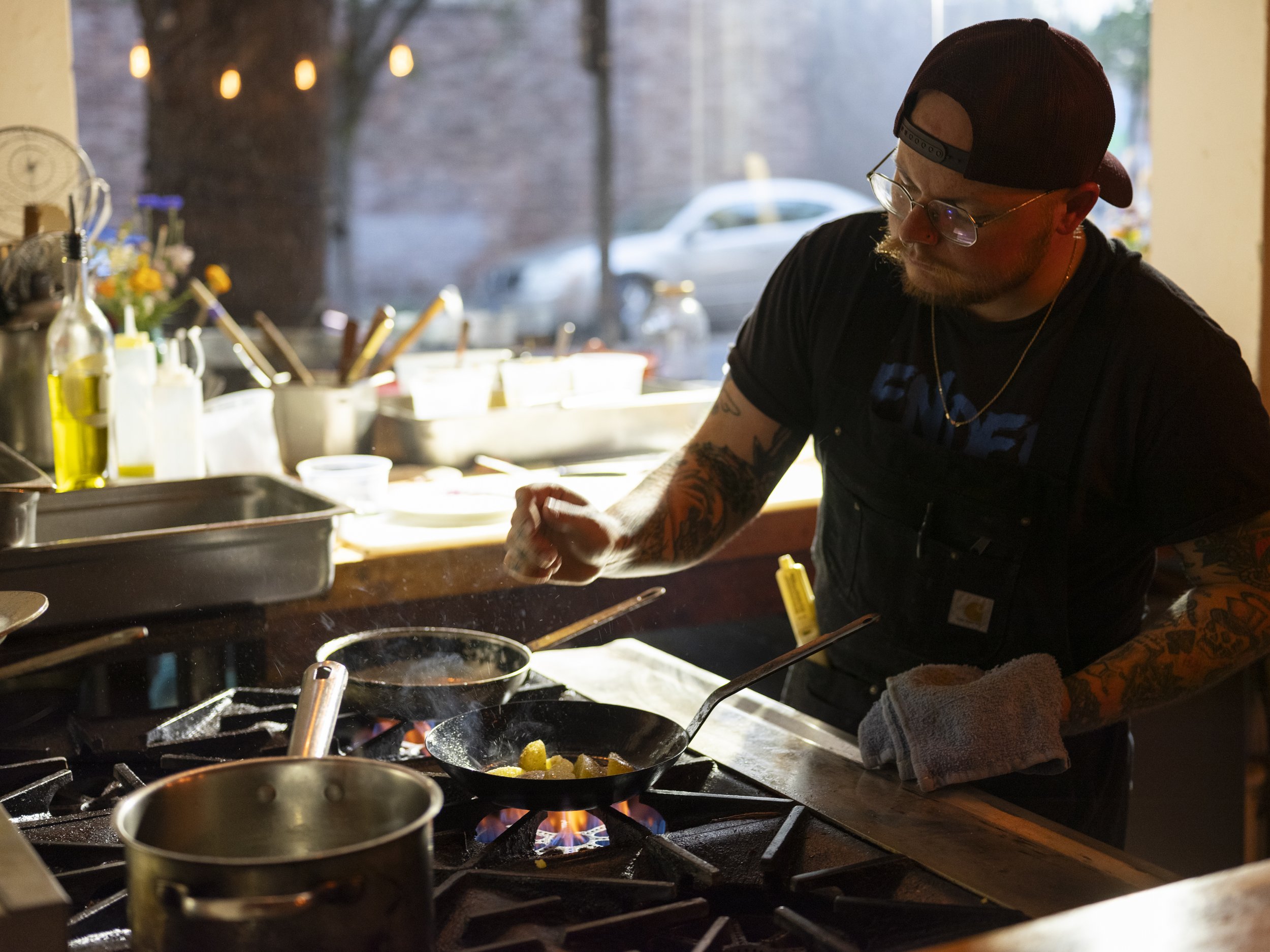 Chef John cooking in a restaurant kitchen, wearing glasses, a backward cap, and a black t-shirt, stirring a skillet with food on a stove.