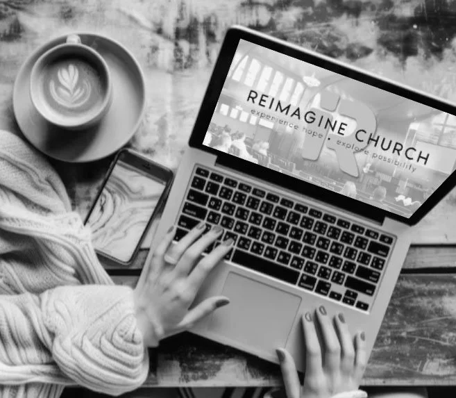 Group of people sitting around tables in a room, with some reading, talking, or looking towards the front, under a wooden ceiling with hanging lights, with a staircase in the background.