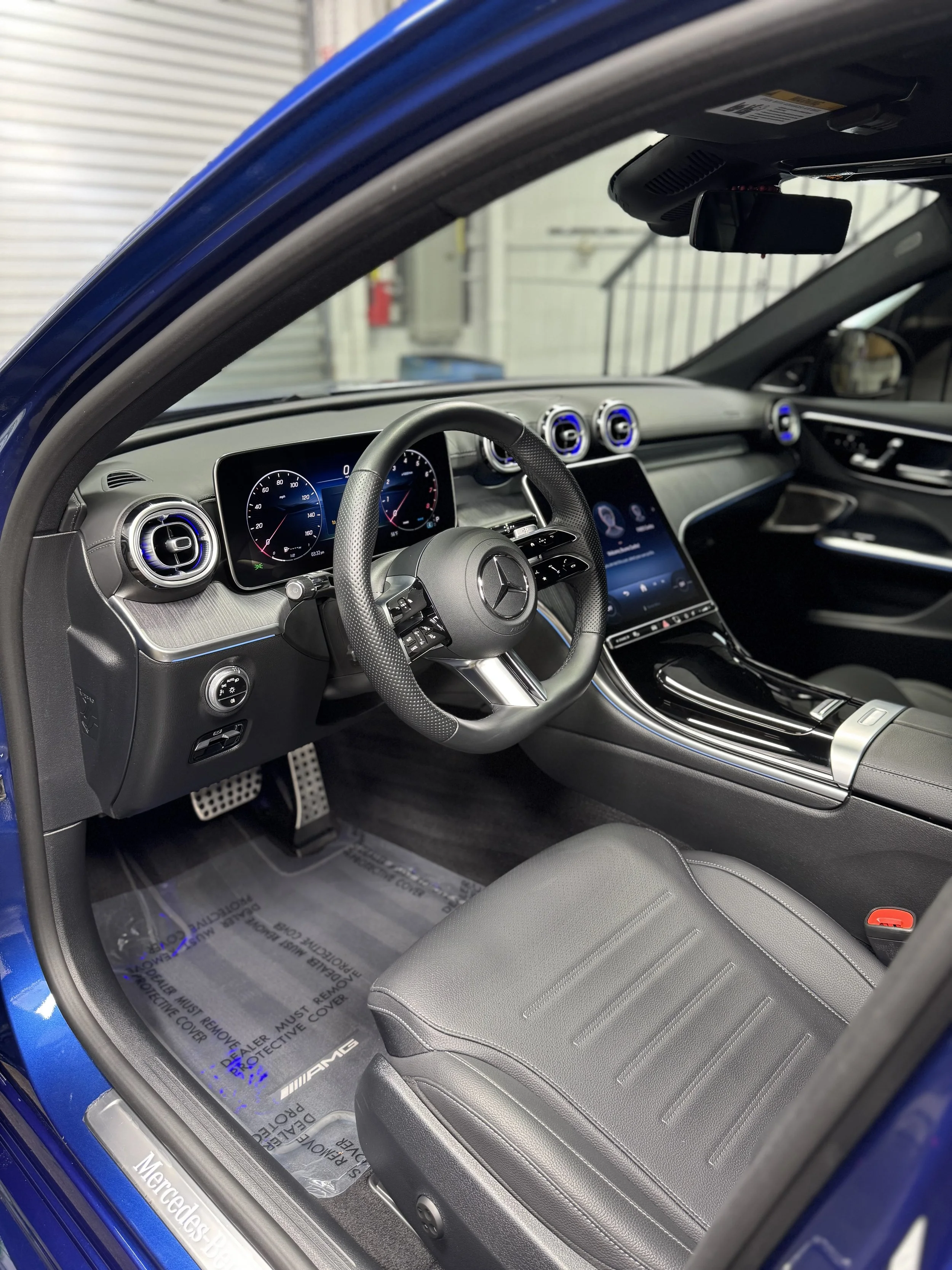 Interior of a blue Mercedes-Benz car showing the dashboard, steering wheel, center console with touchscreen display, and black leather seat.
