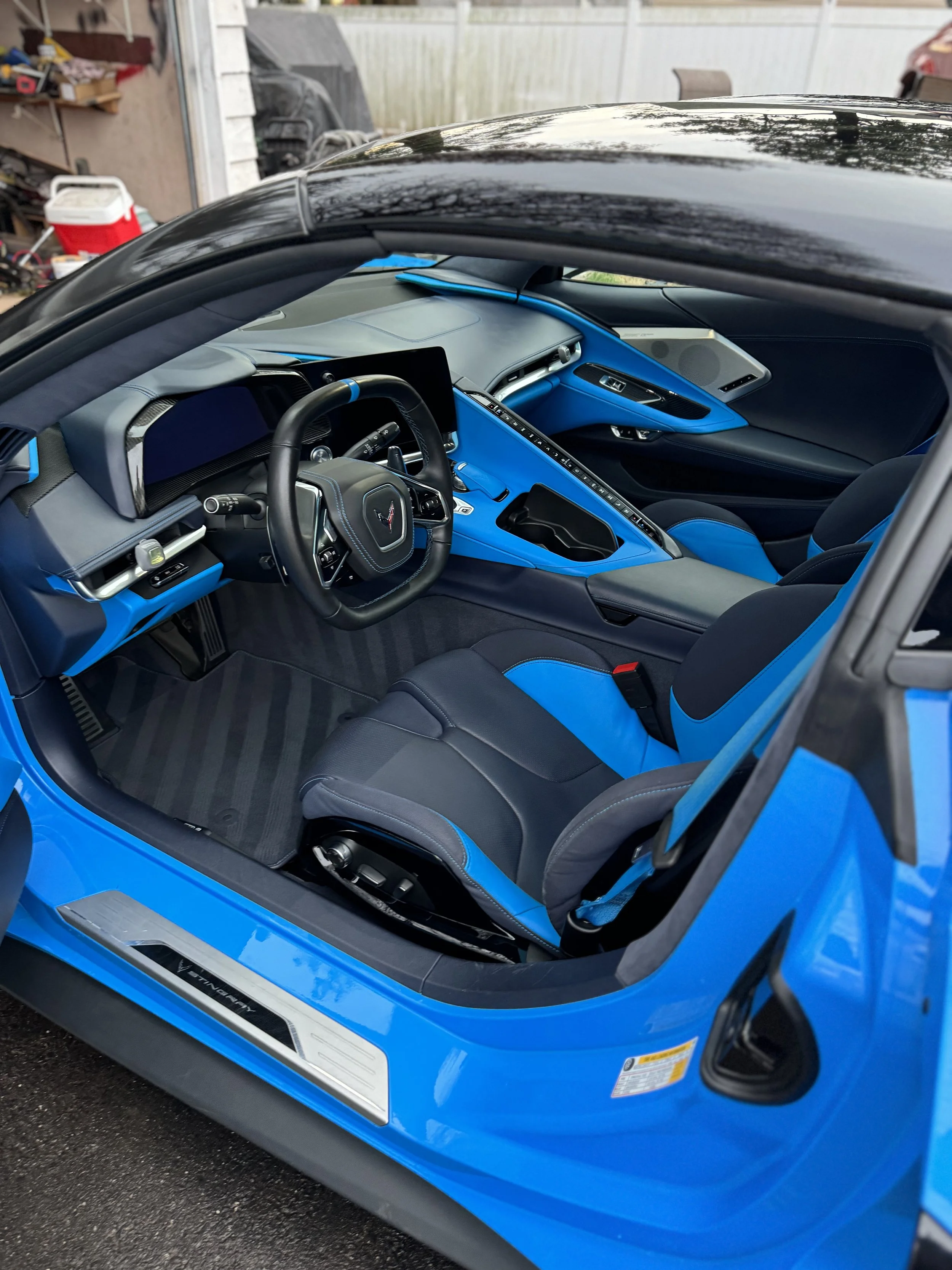 Interior of a blue sports car showing a black and blue racing seat, a black steering wheel with a Corvette emblem, and a dashboard with a digital display and center console.