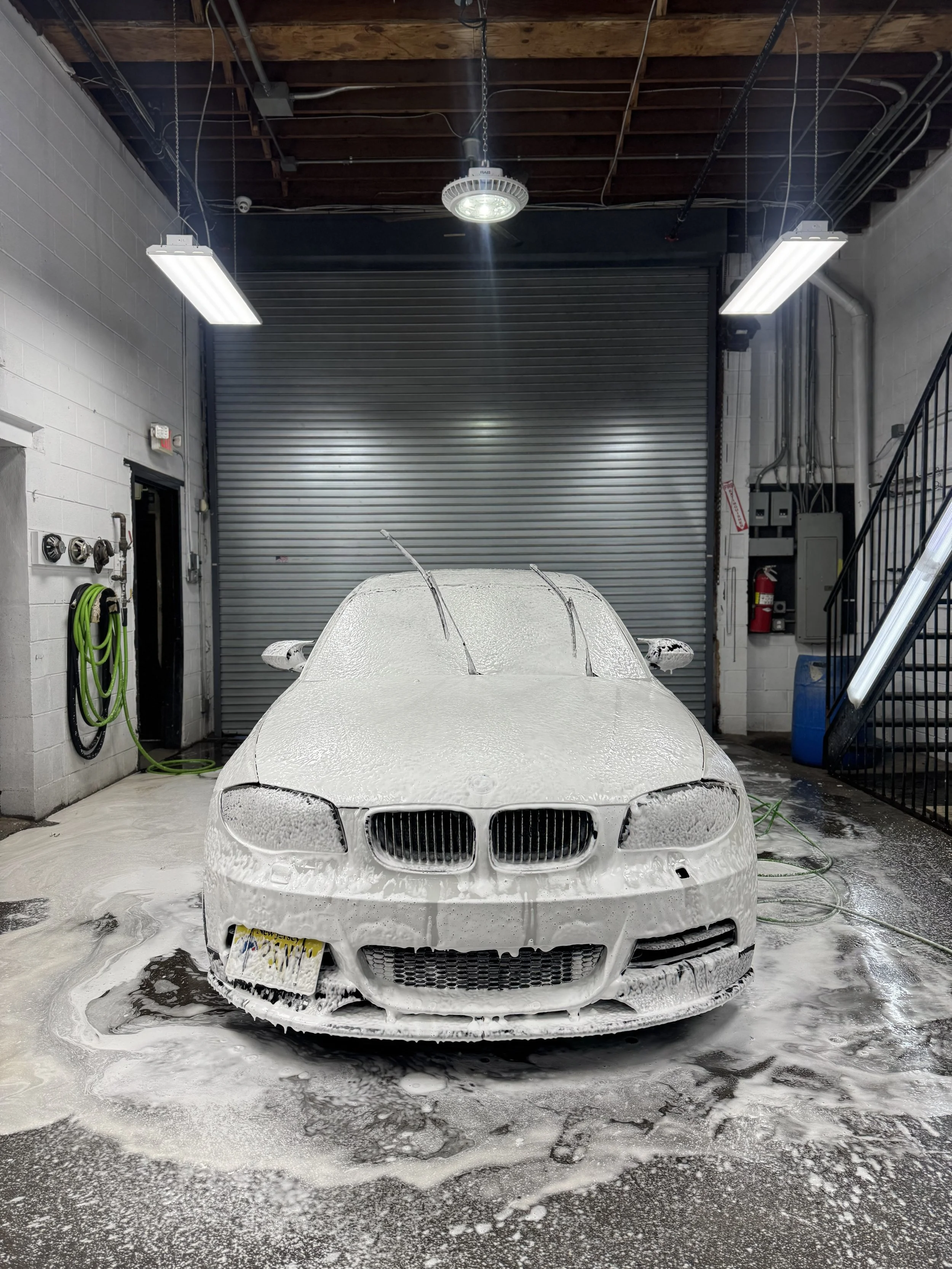 White BMW car covered in soap foam during a car wash in an indoor garage with fluorescent lights and a closed metal garage door.