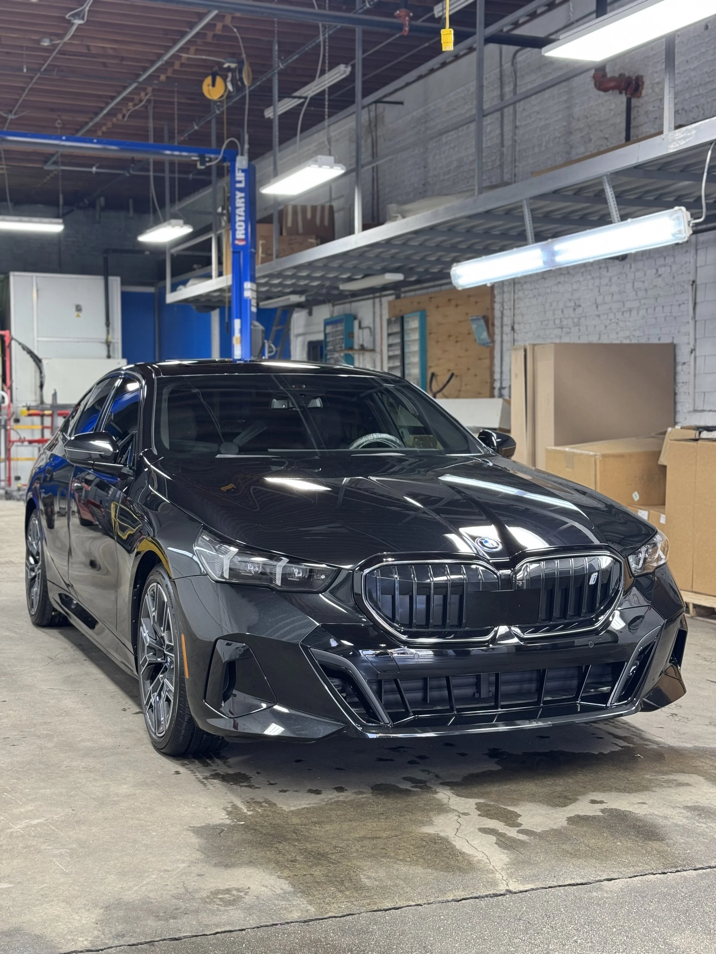 Black sedan car parked indoors in a warehouse or garage setting with some cardboard boxes and equipment in the background.