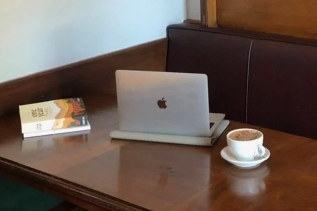 Laptop with Apple logo on wooden table, next to a book and a cup of coffee.