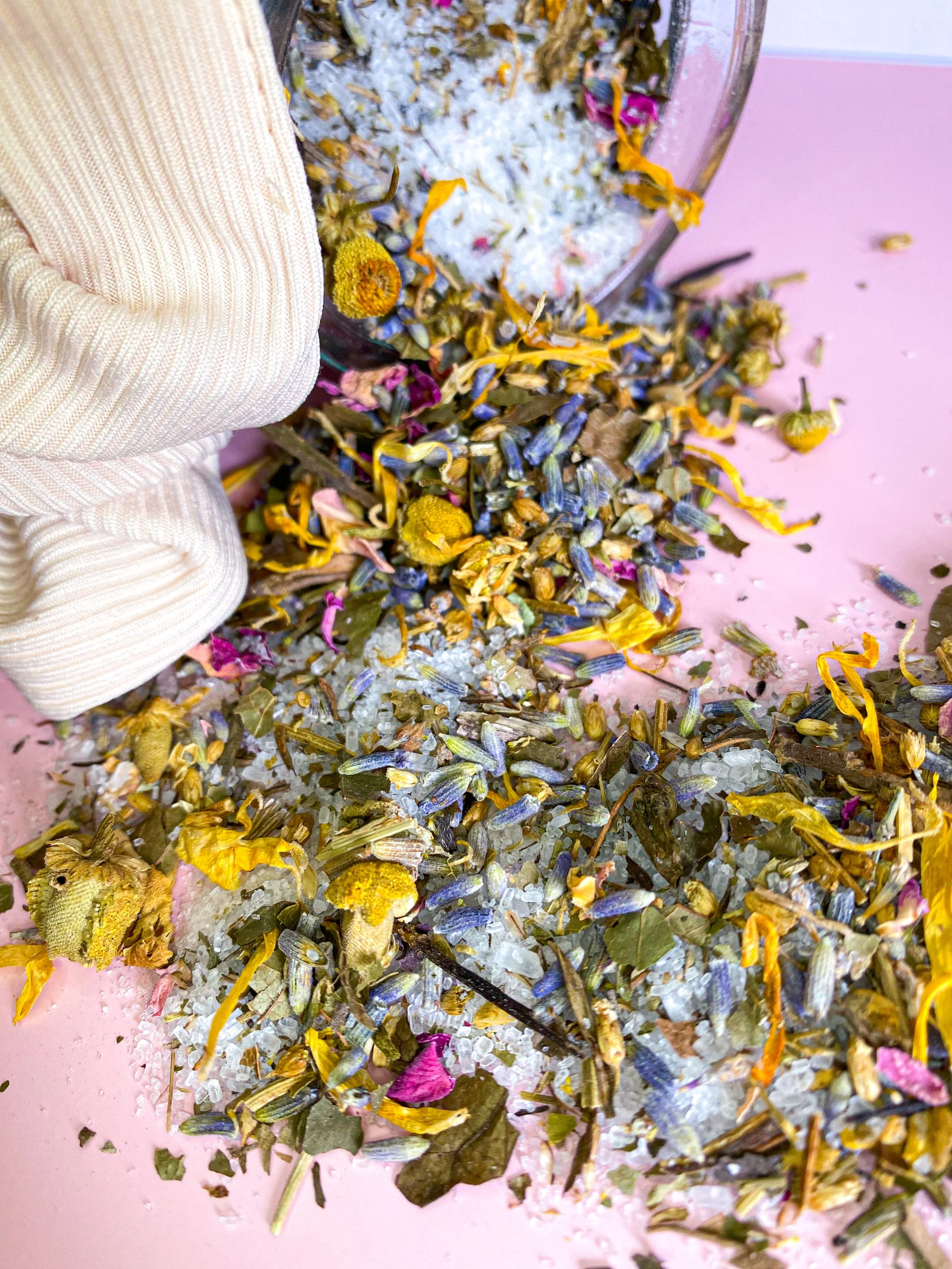 Dried flower petals and herbs spilled on a pink surface with a partial view of a person in a beige knitted glove.