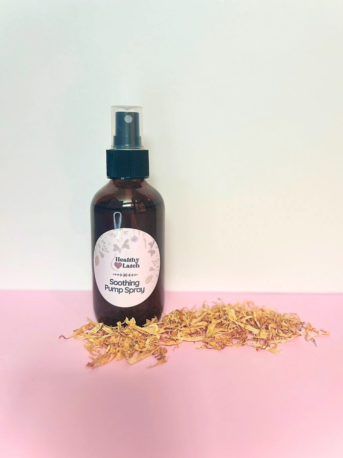 A brown spray bottle labeled 'Healthy Latch Soothing Pump Spray' with dried herbal leaves in front, placed on a pink surface against a white background.