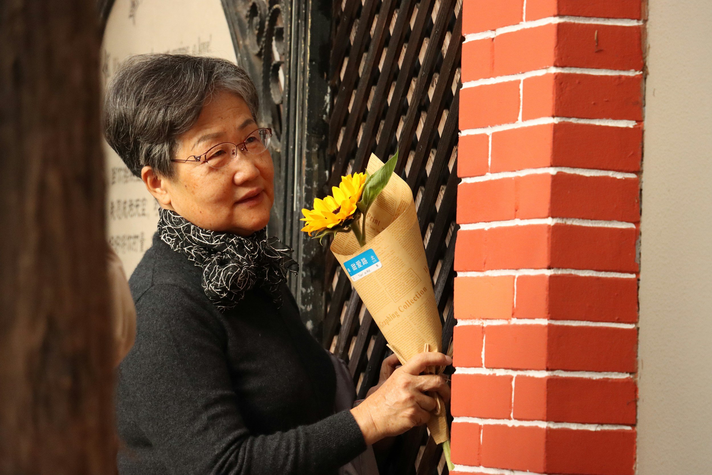 An elderly woman with gray hair and glasses holding a wrapped sunflower bouquet, looking at a brick wall while attending a public event.