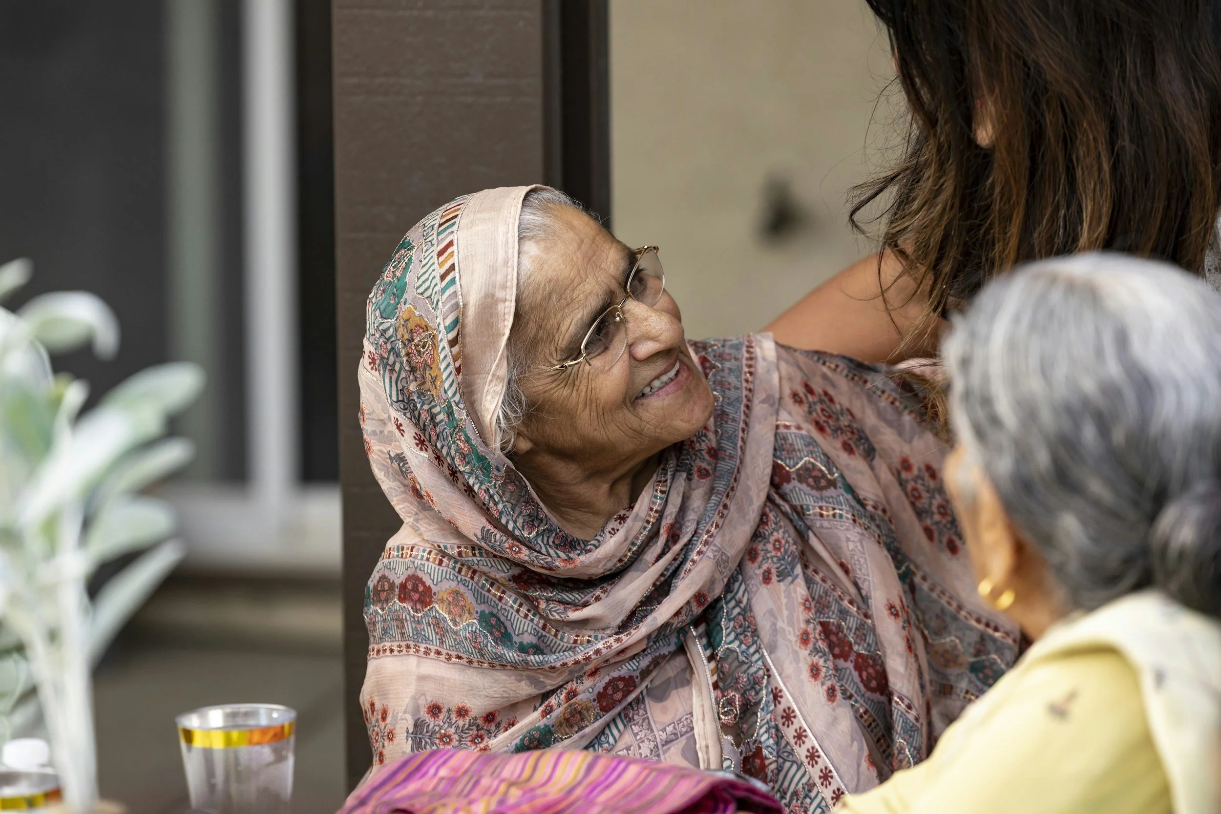 An elderly woman wearing glasses and a colorful patterned headscarf smiling at a person in a restaurant or cafe.
