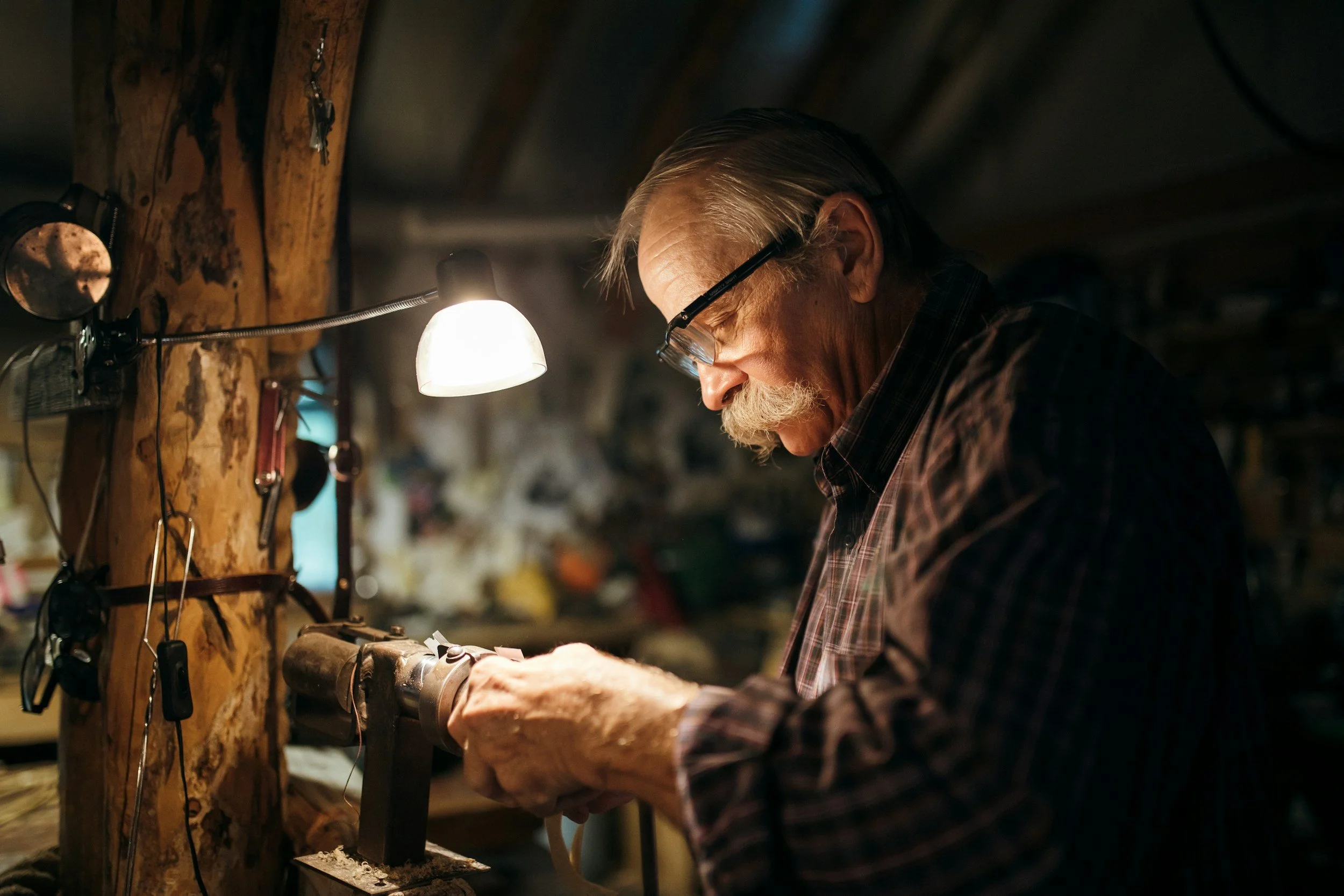 An elderly man with gray hair and a beard working on a woodworking project in a workshop, wearing glasses and checkered shirt, illuminated by a desk lamp.