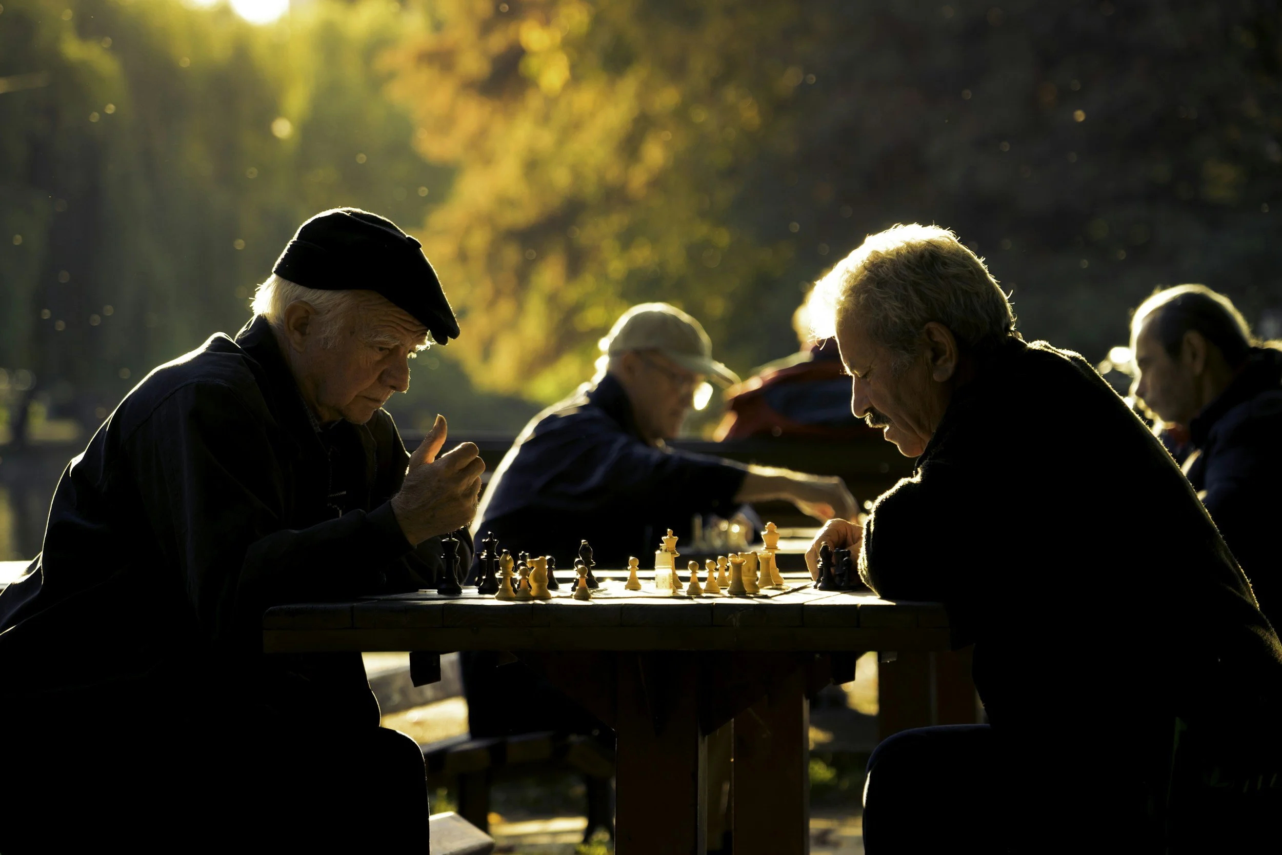 Older men playing chess outdoors at a park during sunset, with warm sunlight filtering through trees in the background.