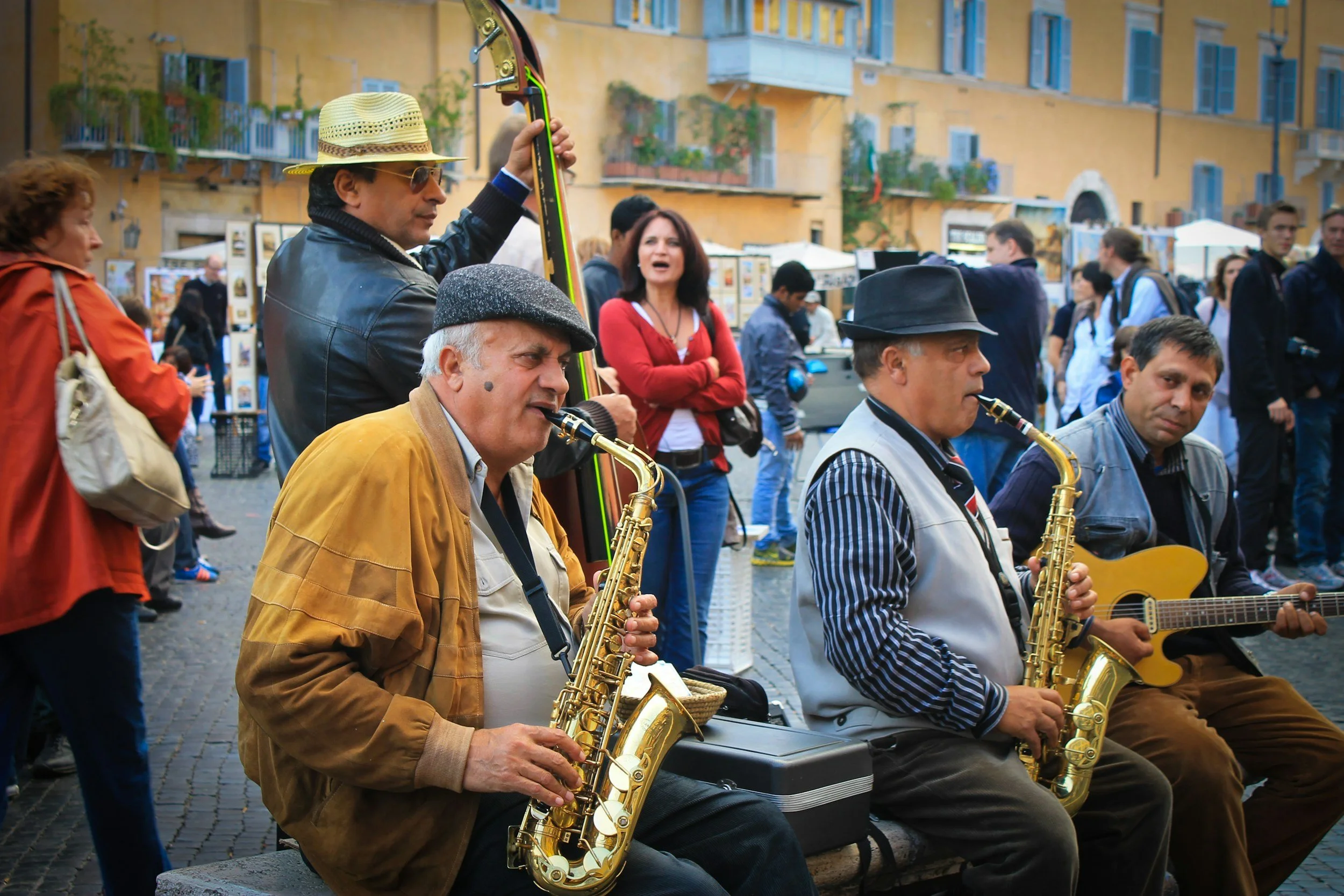 Three musicians playing saxophones and guitar on a cobblestone street during a public event, with people walking and standing in the background near colorful buildings.