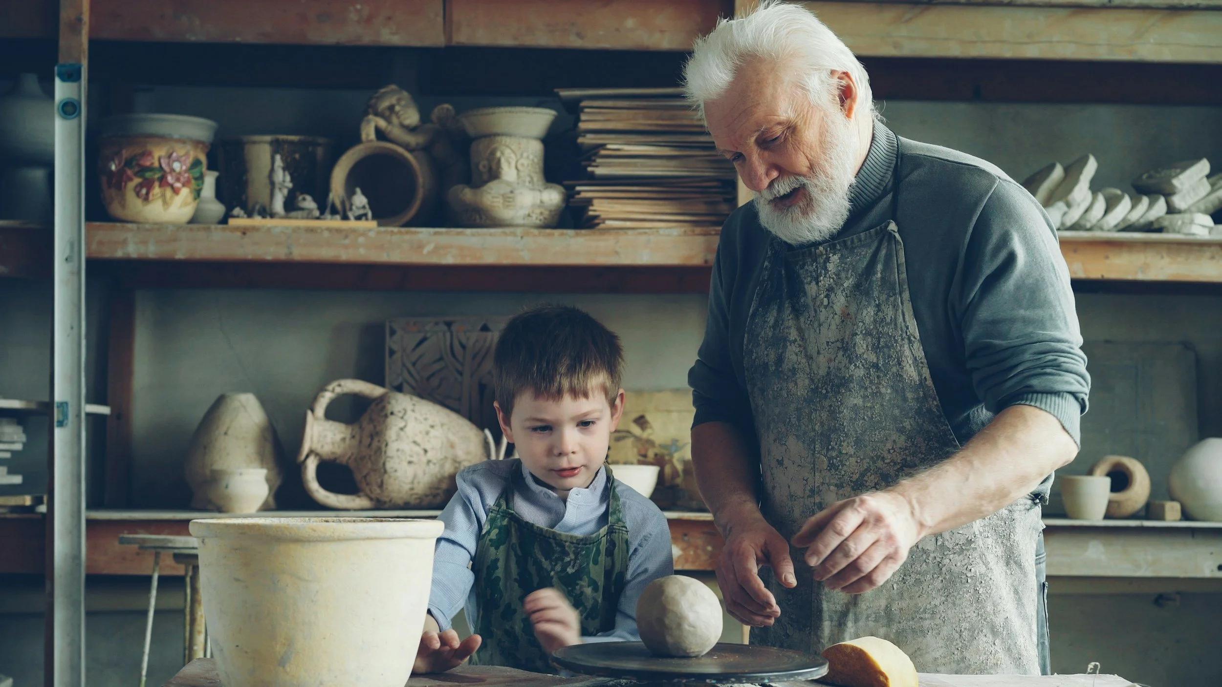 An elderly man and a young boy are working together in a pottery workshop, shaping clay on a pottery wheel.