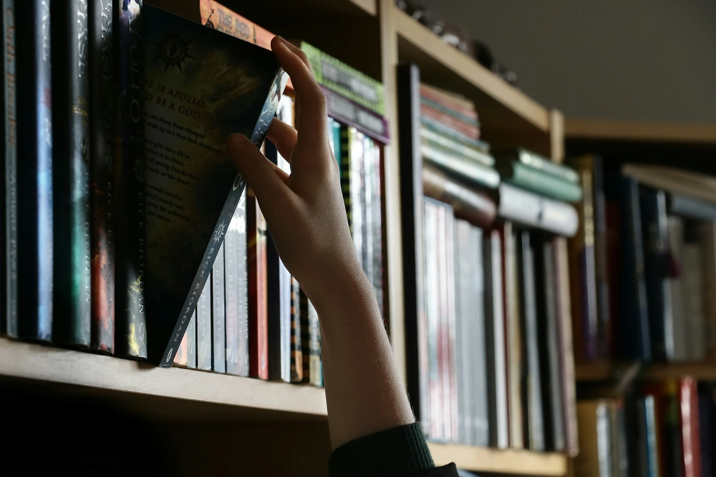 A person shelves a book in a library or bookstore. The person's arm is visible, wearing a black sleeve, and they are placing a book among other books on a wooden shelf filled with books.
