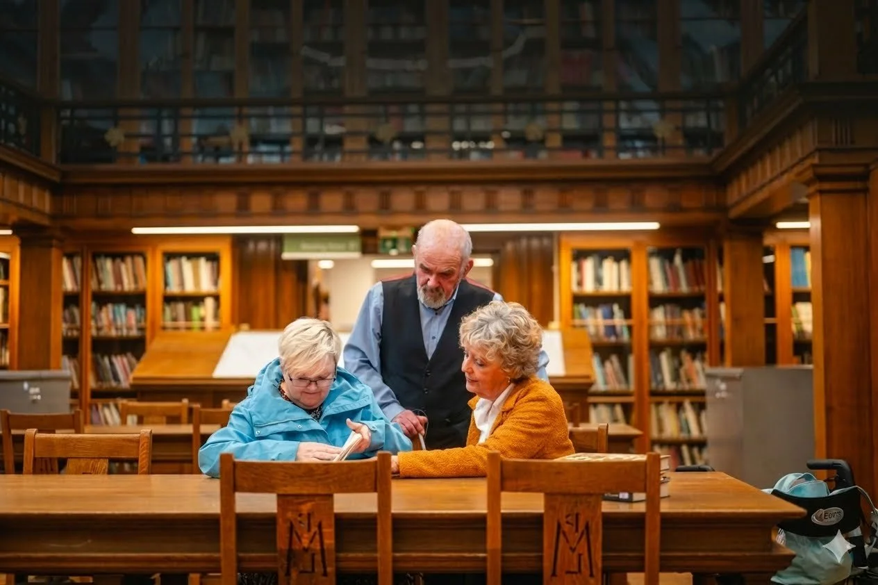 Three elderly individuals, two women and one man, sitting and standing around a table in a library, engaging with a book. The library has wooden bookshelves filled with books and a mezzanine level in the background.