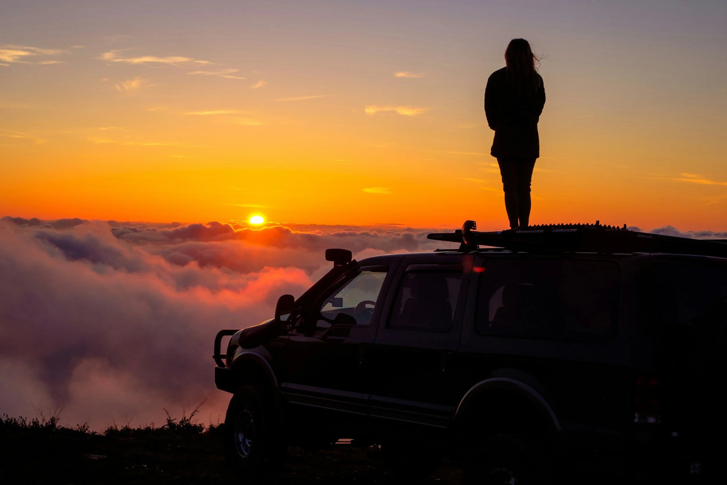 A person standing on top of a dark vehicle, watching a sunset above clouds.