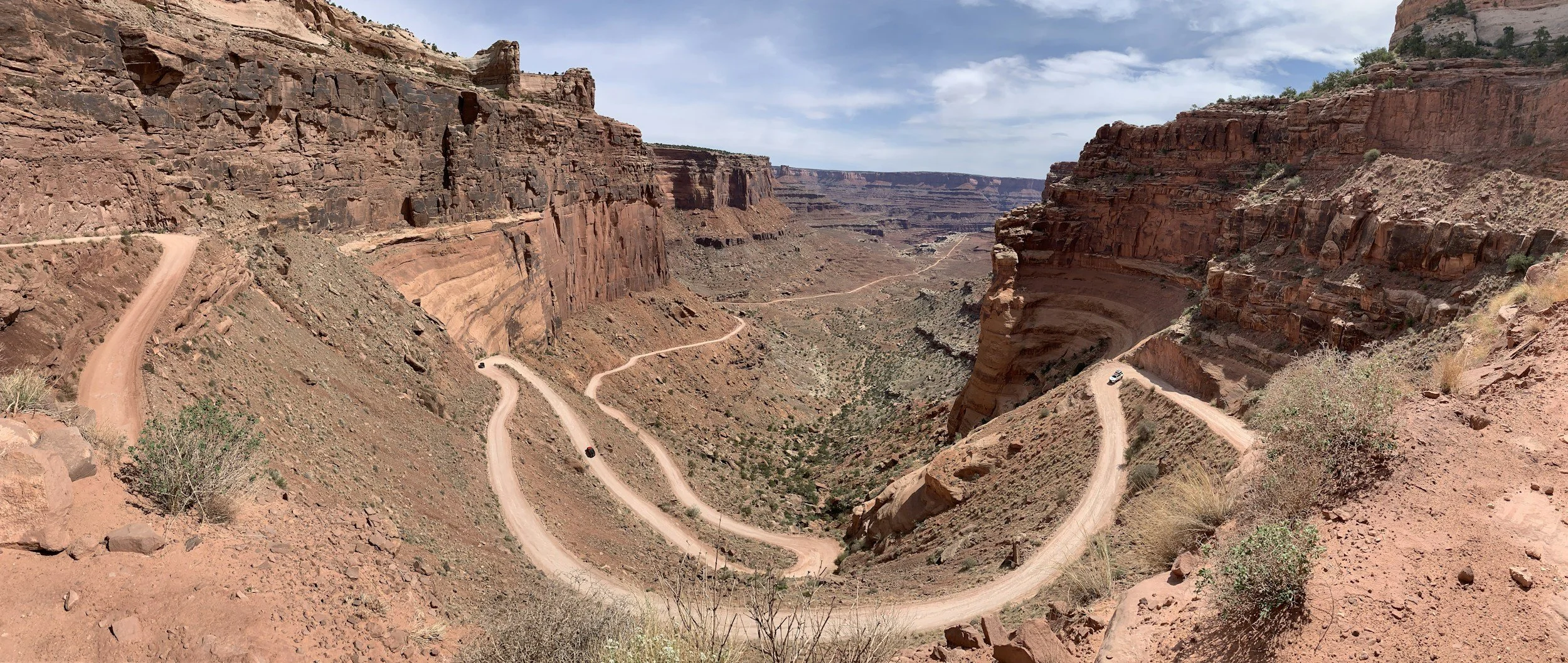 A dusty winding road with switchbacks cuts through a deep canyon with red rock cliffs under a partly cloudy sky.
