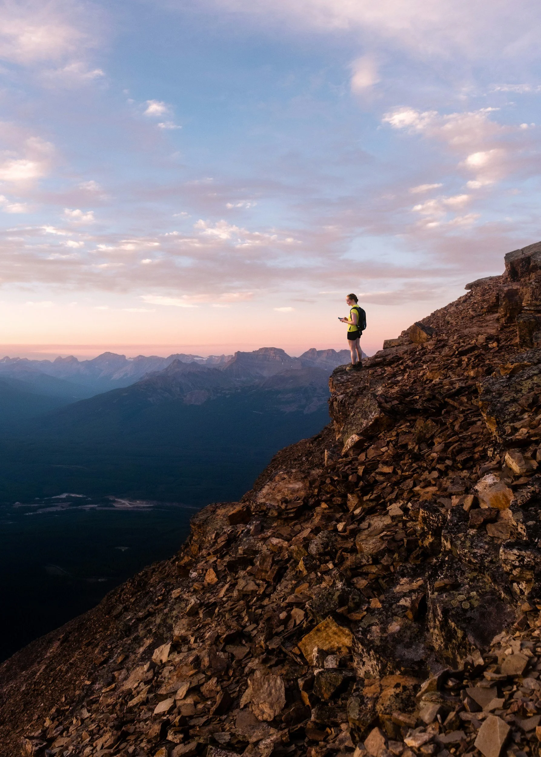 A person in hiking gear standing near the edge of a rocky cliff during sunset or sunrise, overlooking a mountain range.