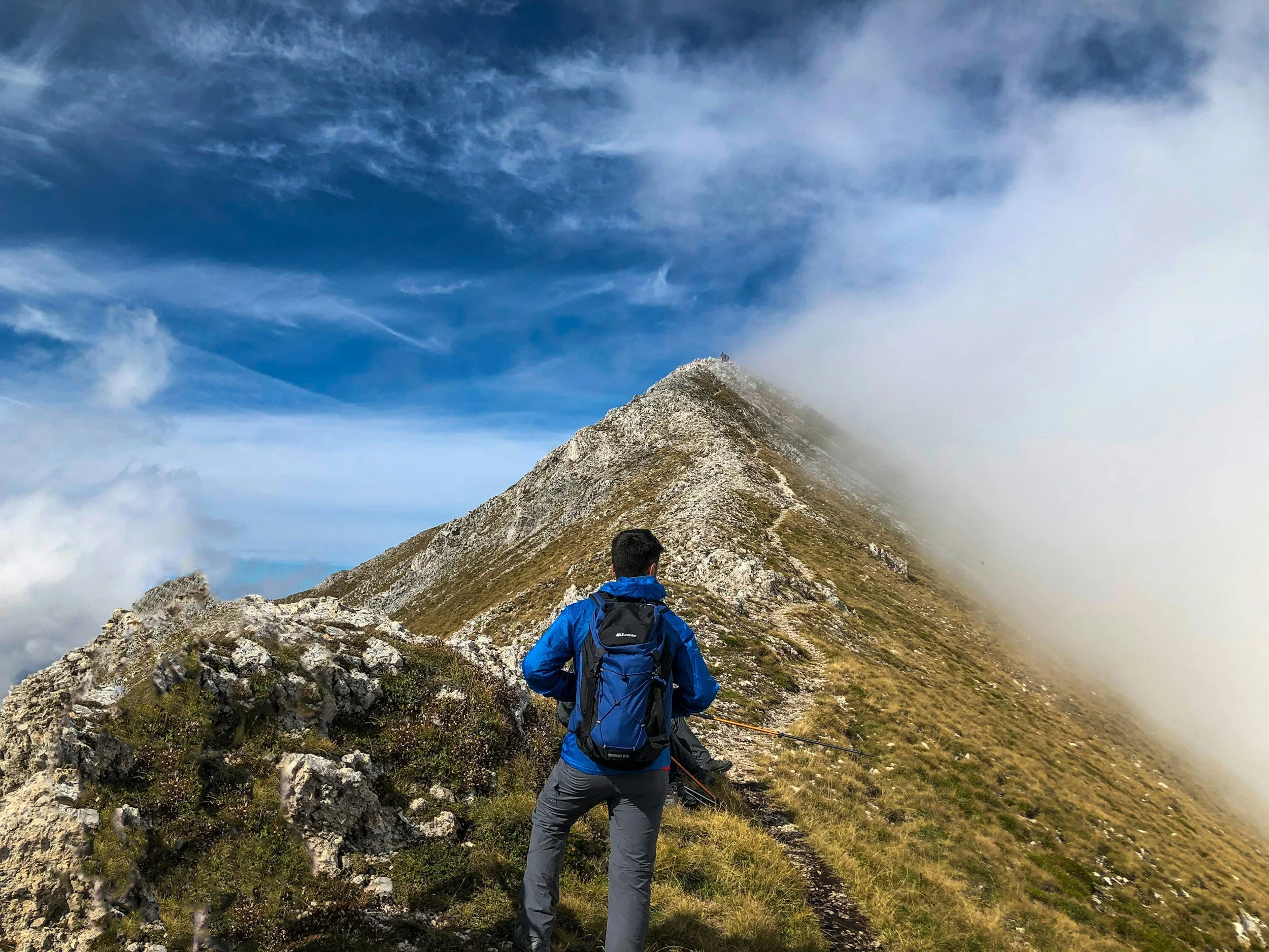 A hiker in a blue jacket and gray pants carrying a backpack, standing on a mountain trail near the summit, surrounded by rocks and grass. The trail continues up a mountain ridge partially covered with mist or clouds, with a partly cloudy sky overhead.