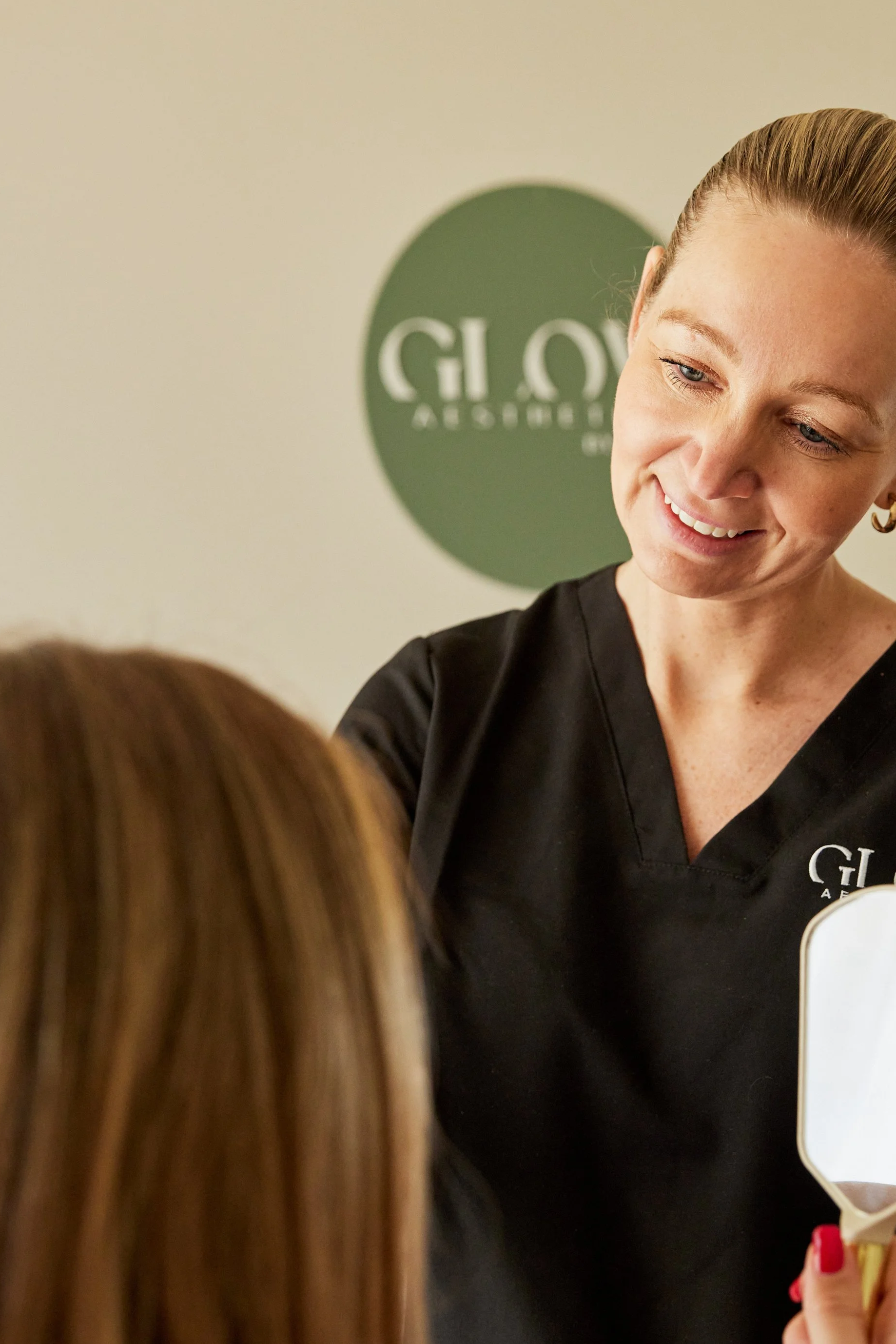 A woman with blonde hair tied back, wearing a black shirt with a logo, smiling and looking at a person with brown hair, while holding a card or paper. In the background, there is a green circular sign with white text that reads 'GIOV'.