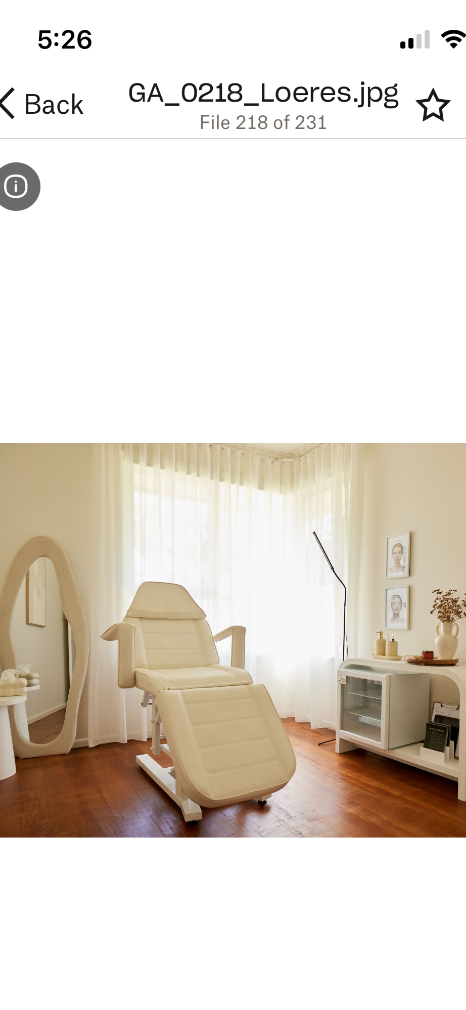 A medical or spa treatment room with a beige recliner chair, side table, and decor like framed pictures, a mirror, and vases with dried flowers, illuminated by natural light from sheer curtains.