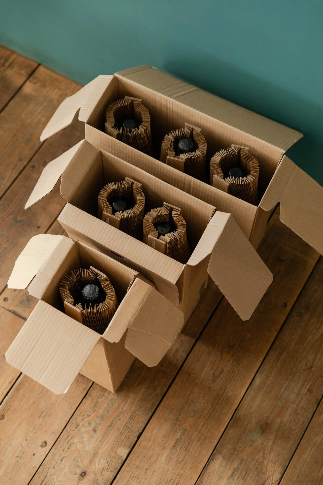 Three open cardboard boxes each containing four wine bottles wrapped in protective packaging, placed on a wooden floor.